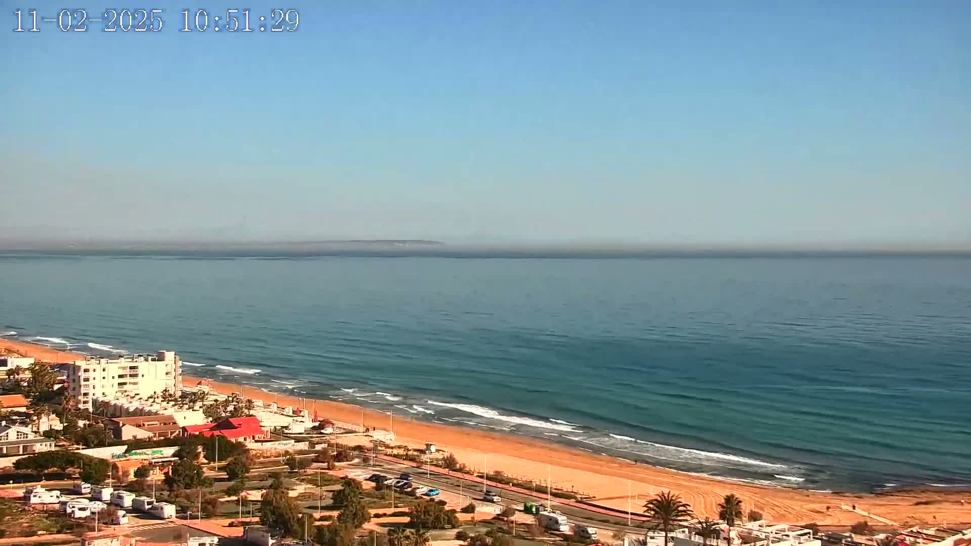 A sandy beach with light waves and a calm ocean under a clear blue sky is visible from an elevated viewpoint, showing buildings and vegetation along the coast.