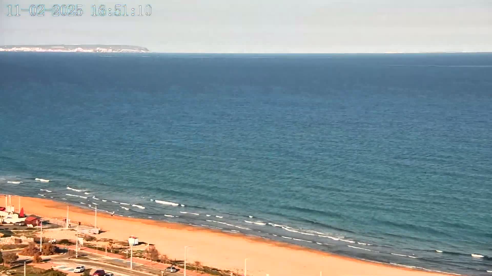 A sandy beach meets a calm, blue ocean under a clear sky, with a distant landmass visible on the horizon.