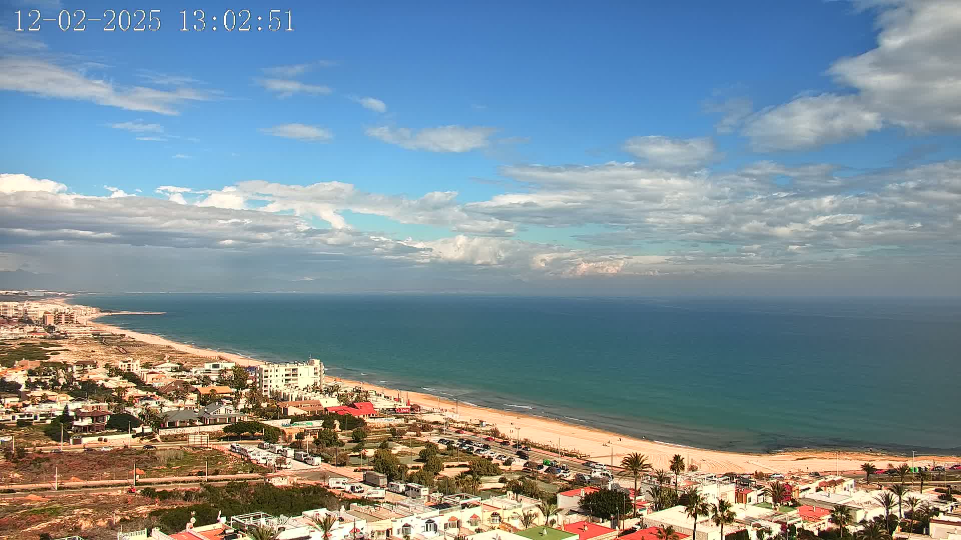 A coastal town with white buildings is situated next to a long, sandy beach and a calm, teal ocean under a partly cloudy sky.