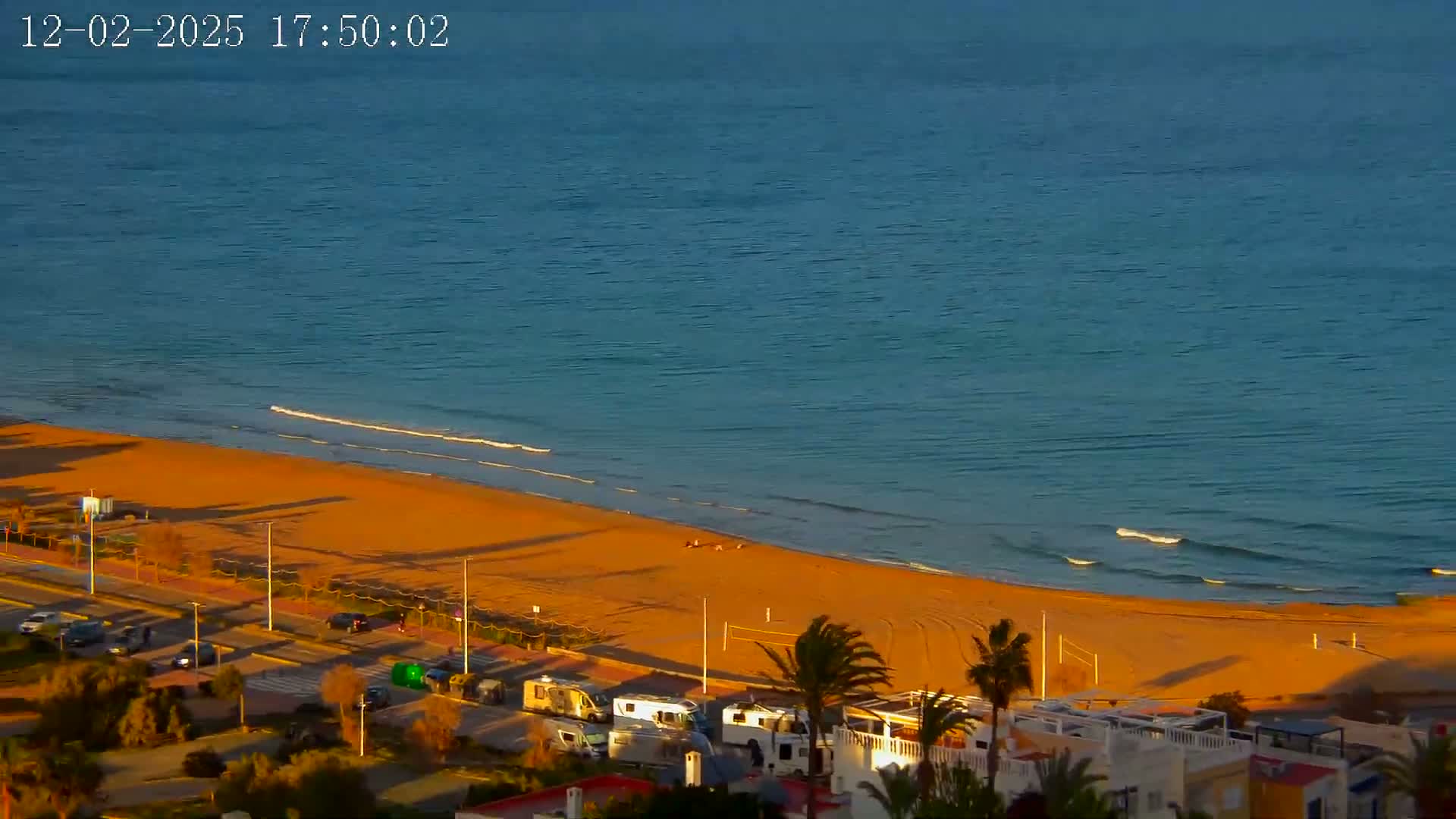 An aerial view shows a sandy beach meeting a calm, blue ocean under a clear sky, with several RVs parked near buildings behind the beach.