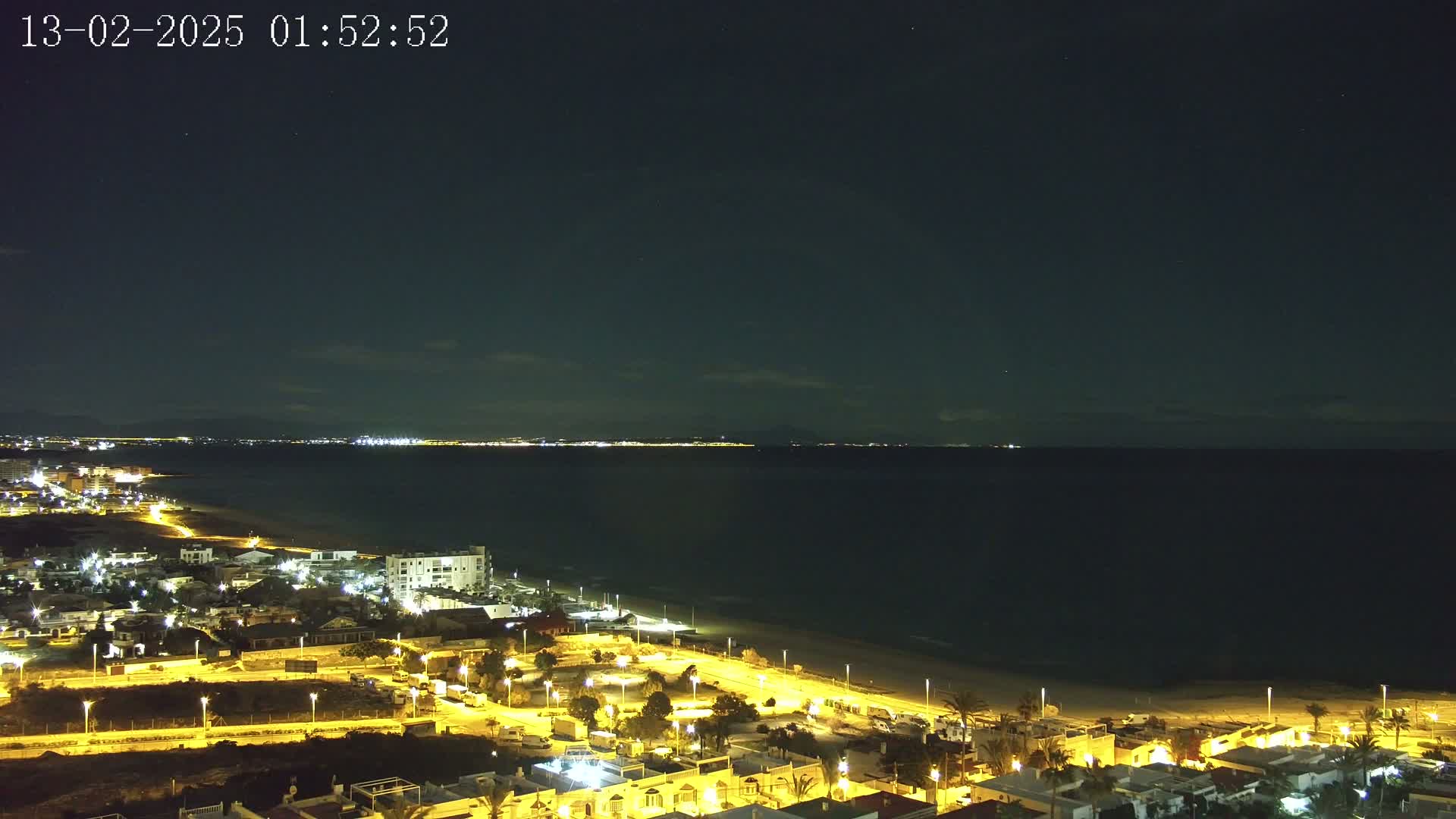 A nighttime coastal town is brightly lit, overlooking a dark sea under a clear, starry sky.