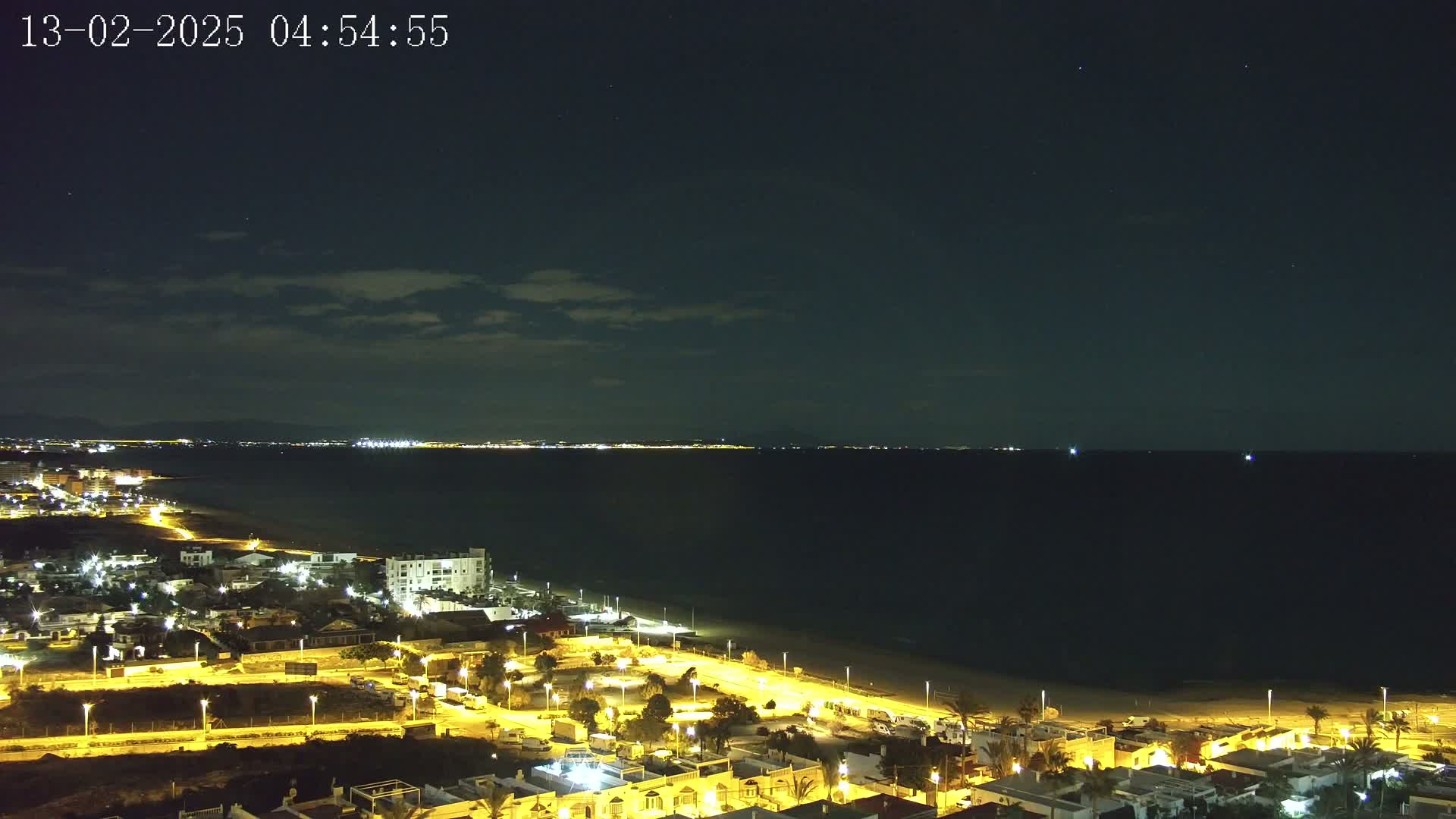 A nighttime coastal view shows a dark ocean under a mostly clear sky, with a brightly lit town on the shore extending to a distant, faintly lit coastline.