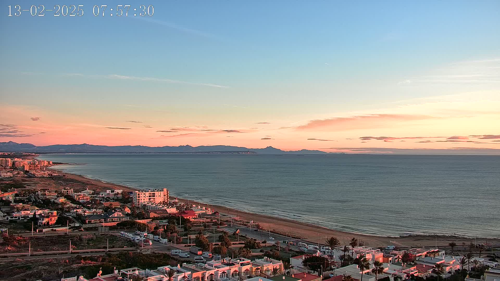 An elevated view of a coastal town at sunset, showing a long sandy beach, calm ocean, and distant mountains under a pale pink and blue sky.