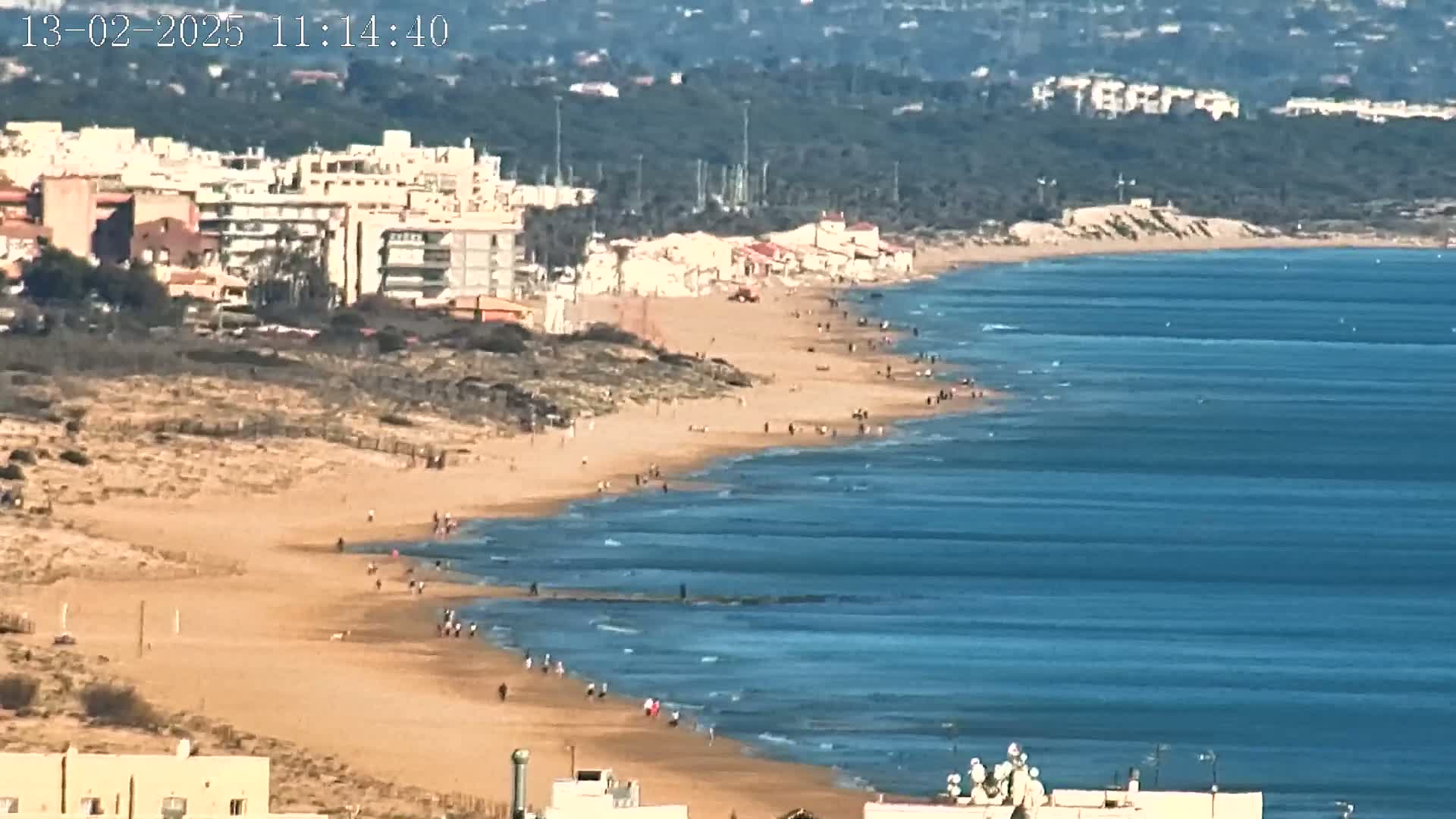A long sandy beach stretches along a calm, blue ocean under a clear sky, with buildings and vegetation visible in the background.