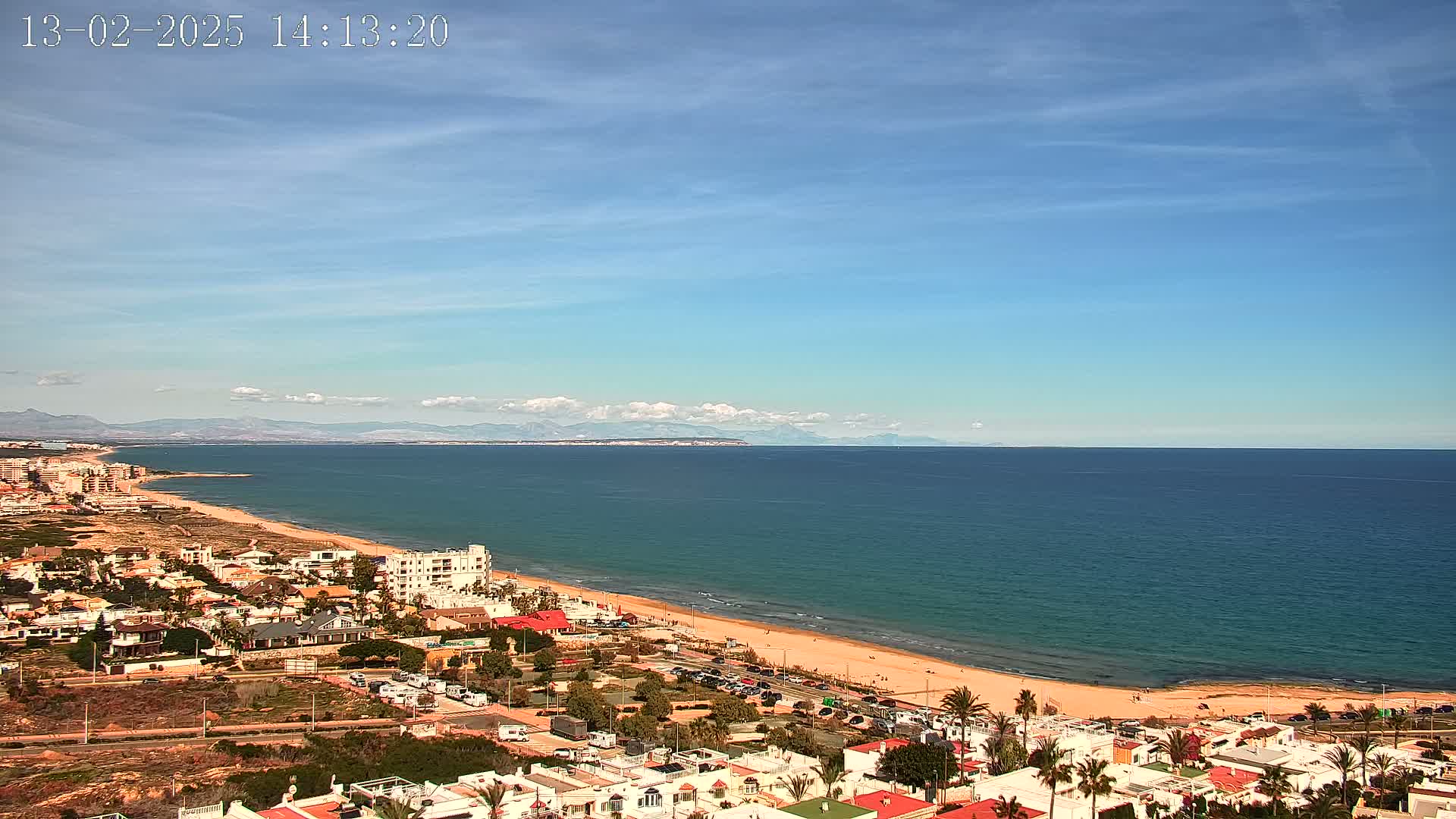 A coastal town with a sandy beach and calm ocean under a mostly clear, bright sky with distant mountains visible on the horizon.
