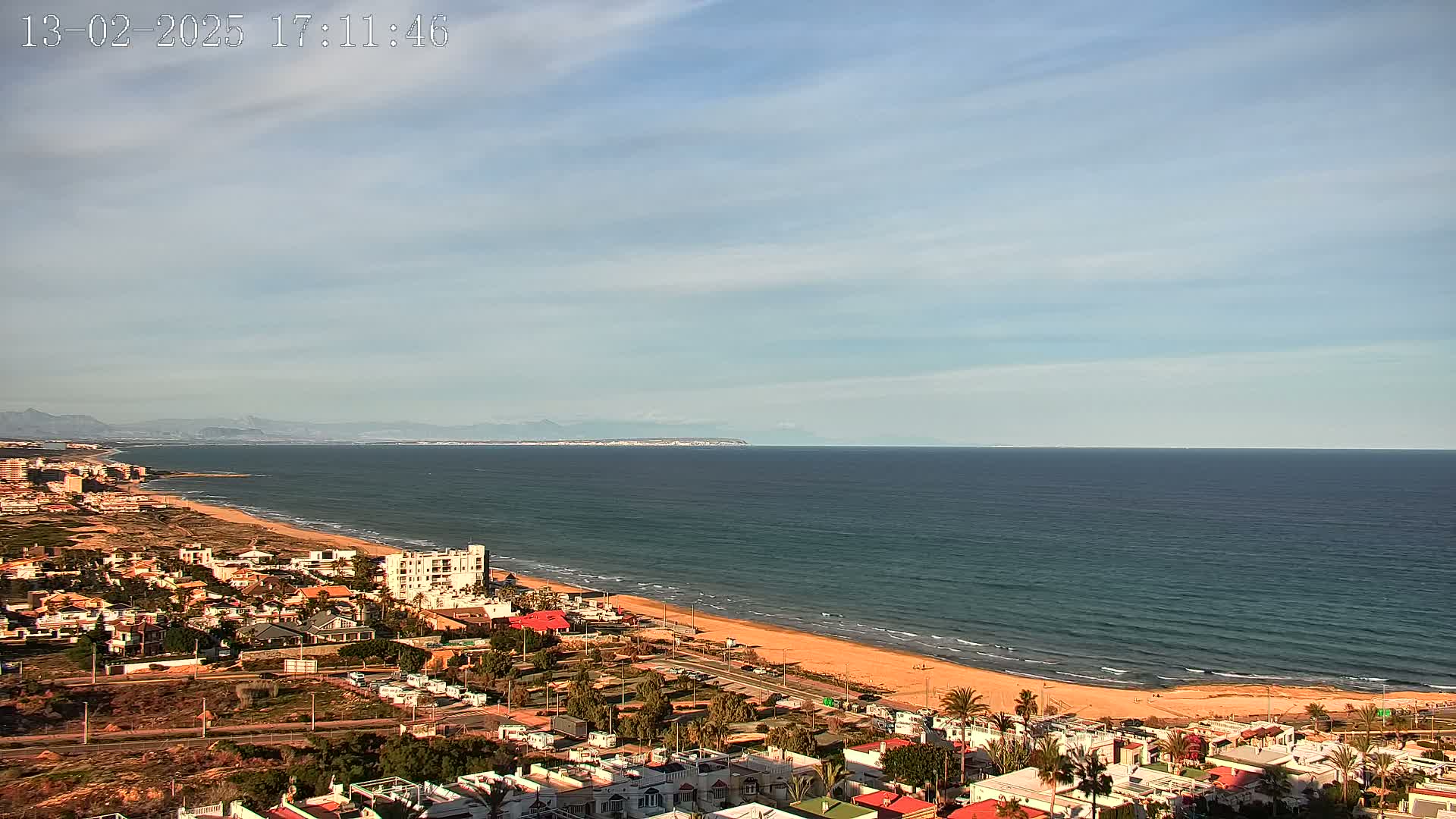 A coastal town with a sandy beach and calm ocean under a mostly clear sky stretches towards distant mountains.