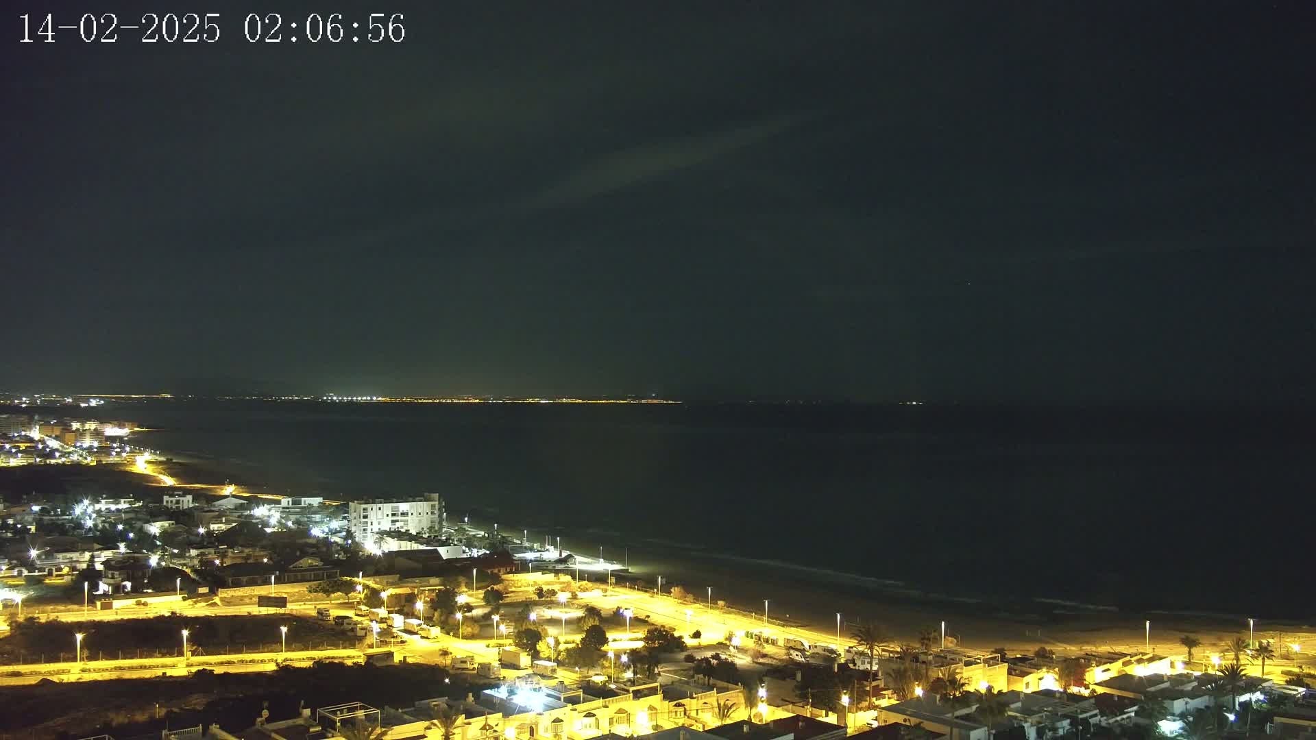 A nighttime coastal town with illuminated buildings and streets is situated next to a dark ocean under a mostly clear sky.
