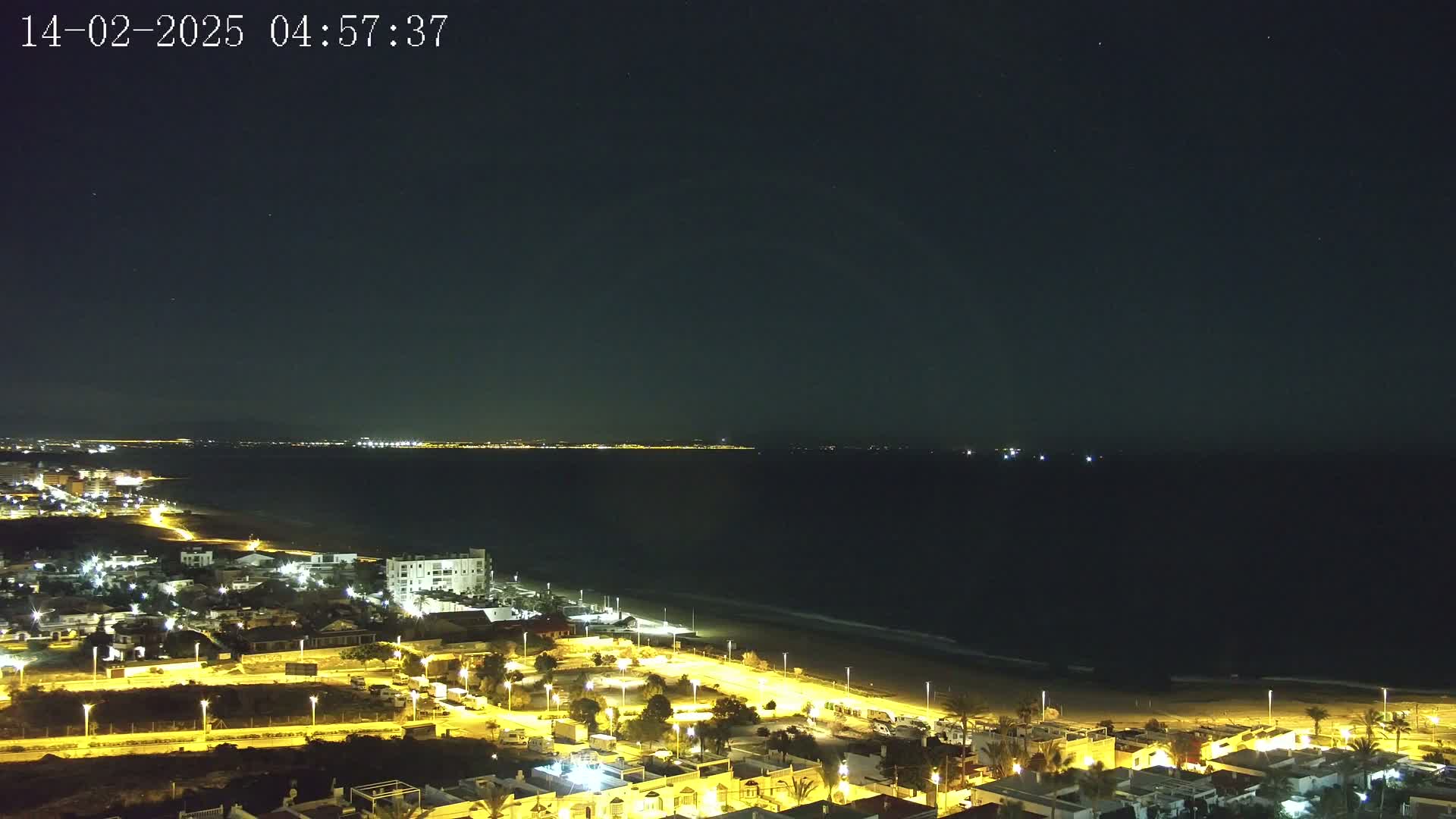 A nighttime, aerial view shows a coastal town illuminated with streetlights, a dark ocean with a sandy beach, and a distant city skyline under a clear, starry sky.