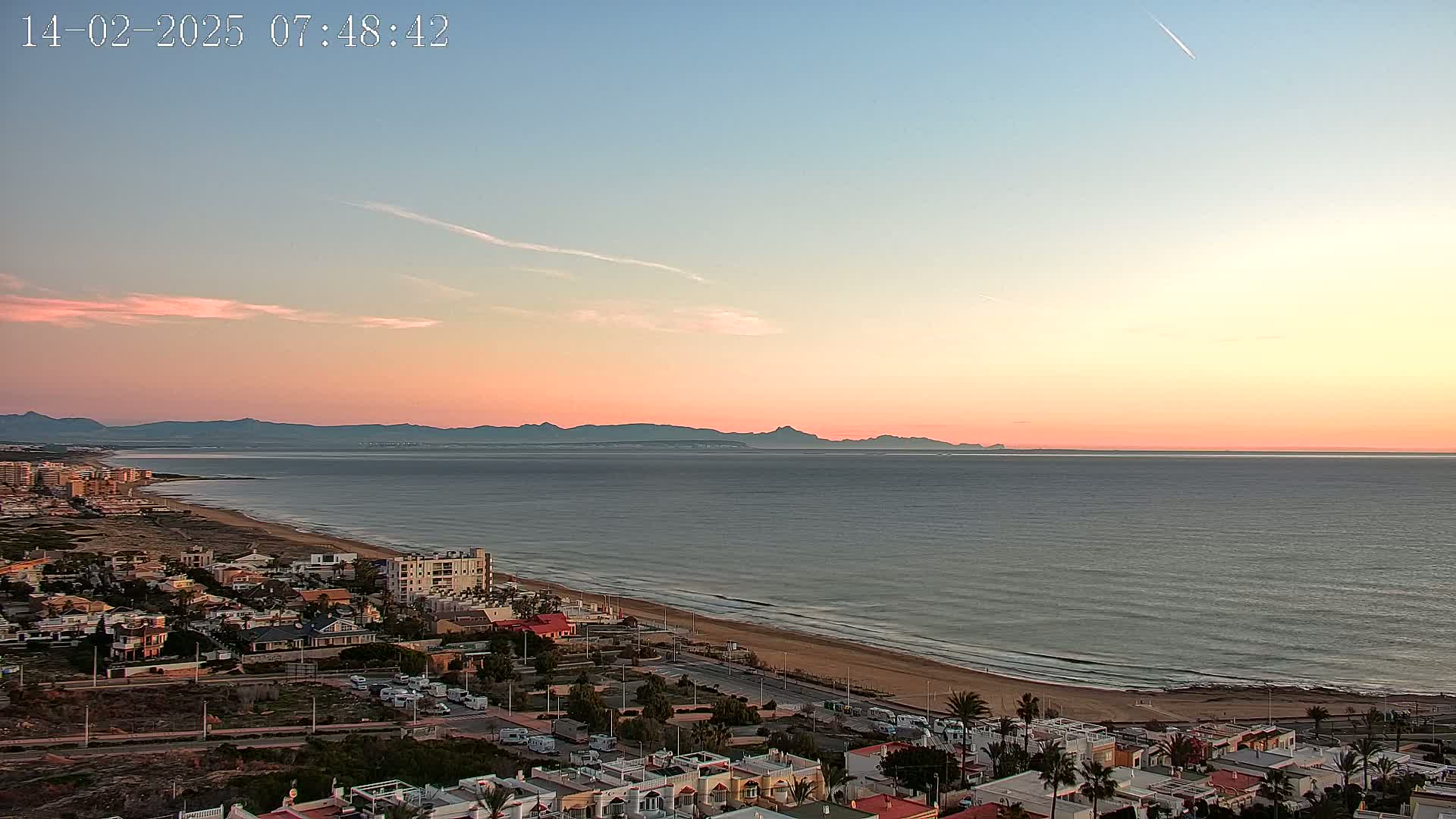 A coastal town with a sandy beach and calm ocean is viewed from above at sunset under a mostly clear sky.