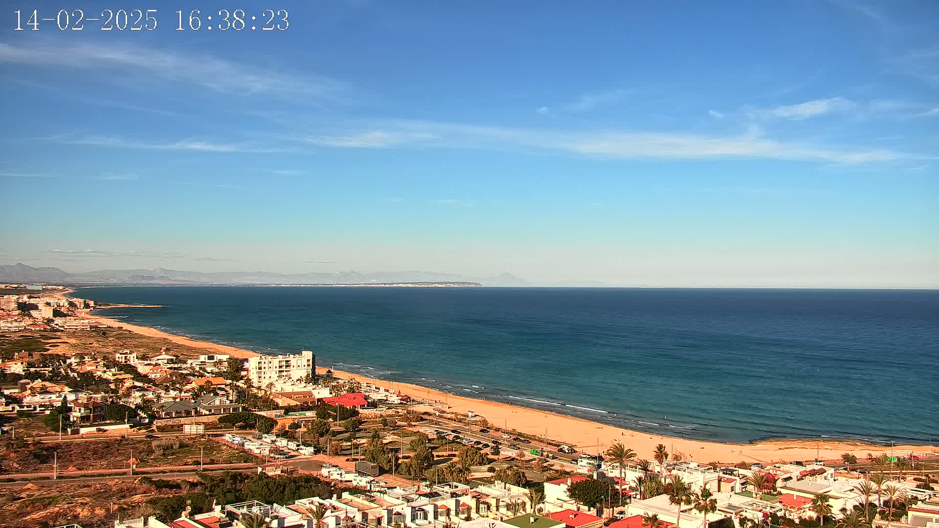 A coastal town with low-rise buildings stretches along a sandy beach and a calm, blue ocean under a clear blue sky.