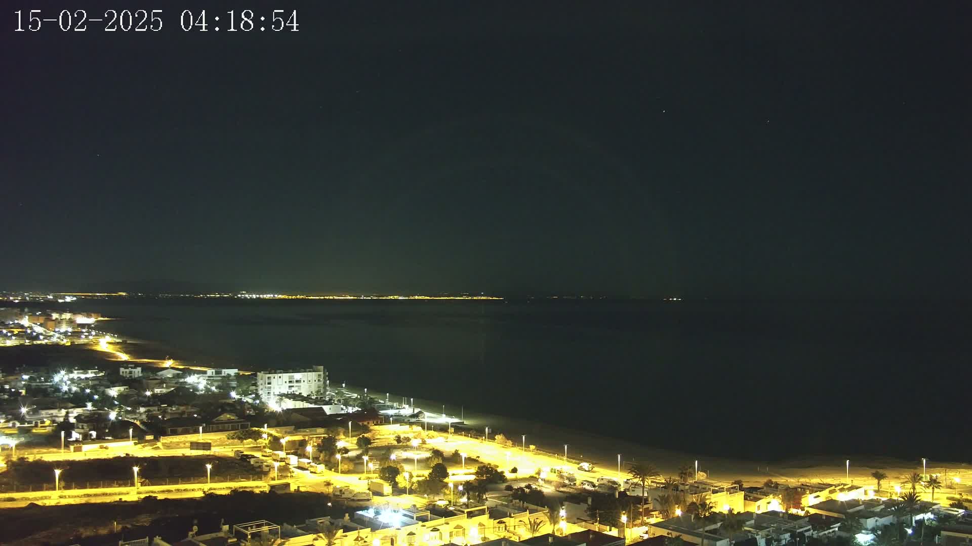 A nighttime, high-angle view shows a coastal town illuminated by streetlights, a dark beach, and a calm, dark body of water extending to a dimly lit horizon under a clear, starlit sky.
