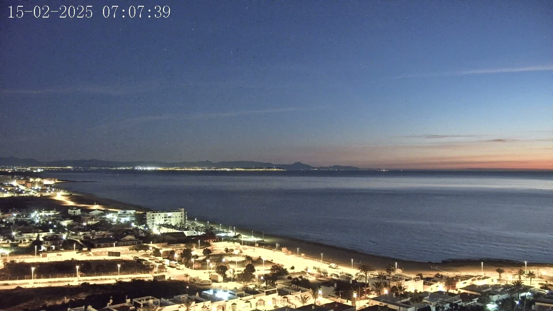 A coastal town at night, illuminated by streetlights, overlooks a calm sea under a twilight sky.