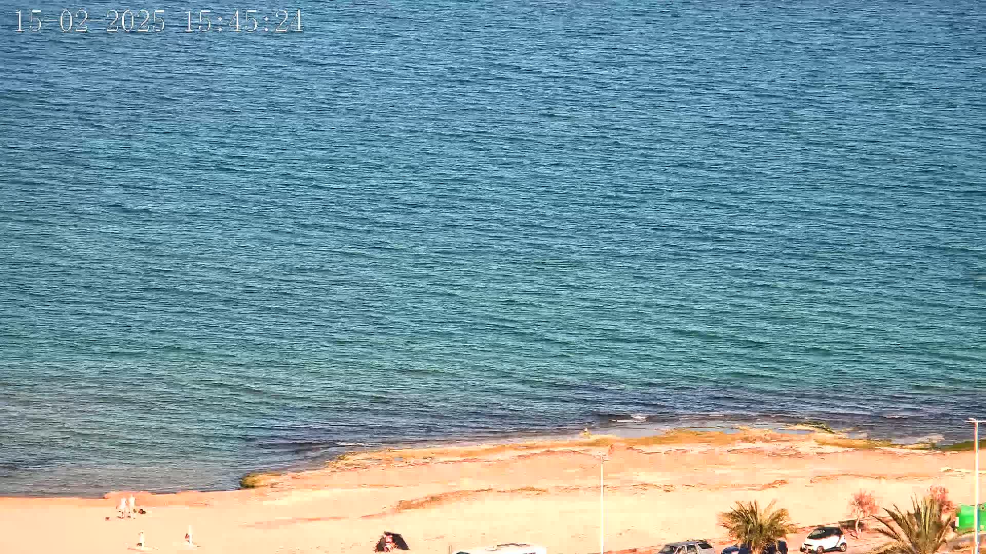 A mostly calm, blue ocean meets a sandy beach with a few people and cars visible.