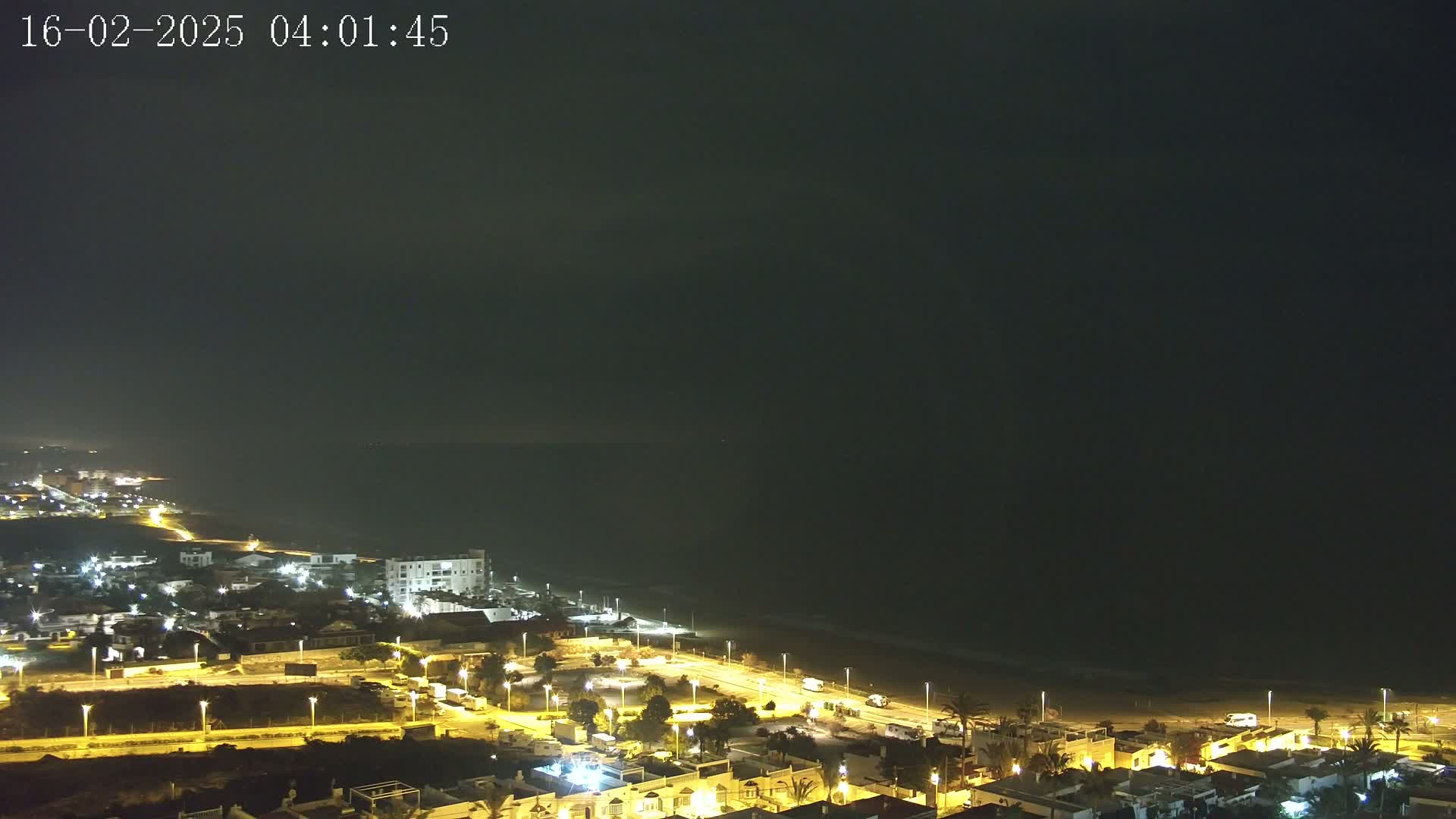 A nighttime coastal town illuminated with streetlights stretches along a dark, sandy beach under a mostly clear, dark sky.