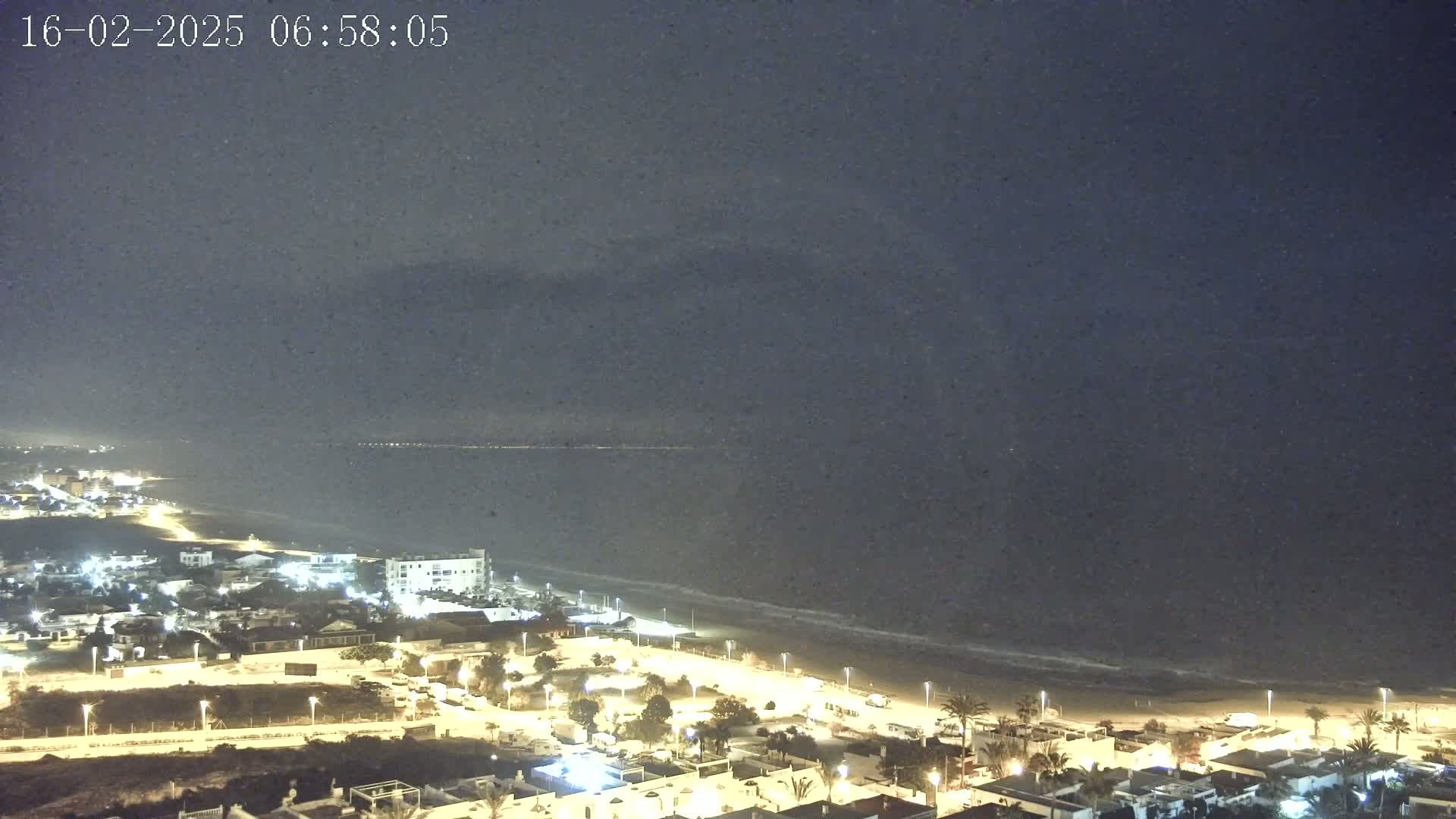 A nighttime view shows a dark ocean meeting a sandy beach, with a coastal town illuminated by streetlights under a slightly hazy sky.