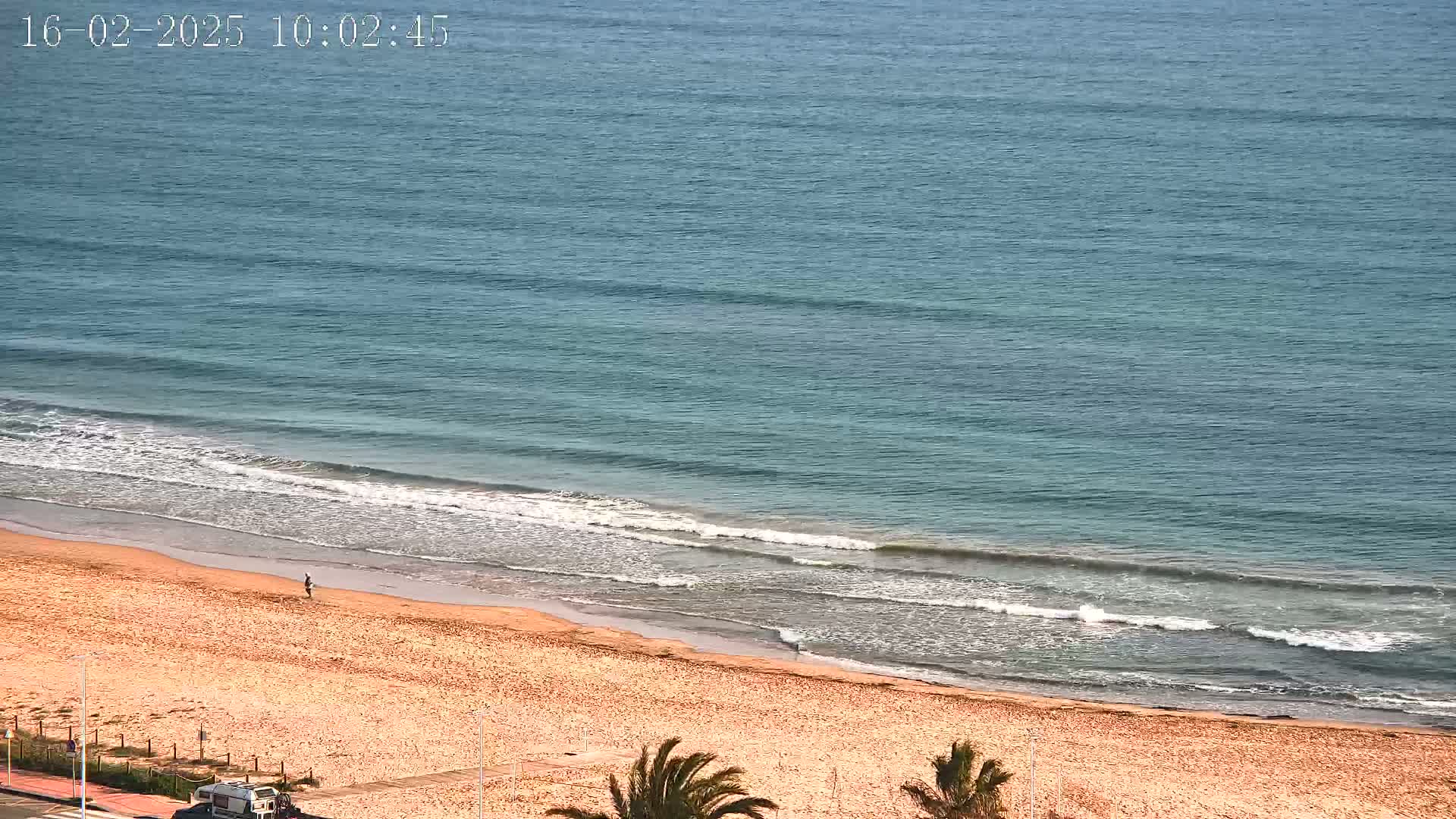 A sandy beach with a lone person walking on it and gentle waves lapping the shore on a sunny day.