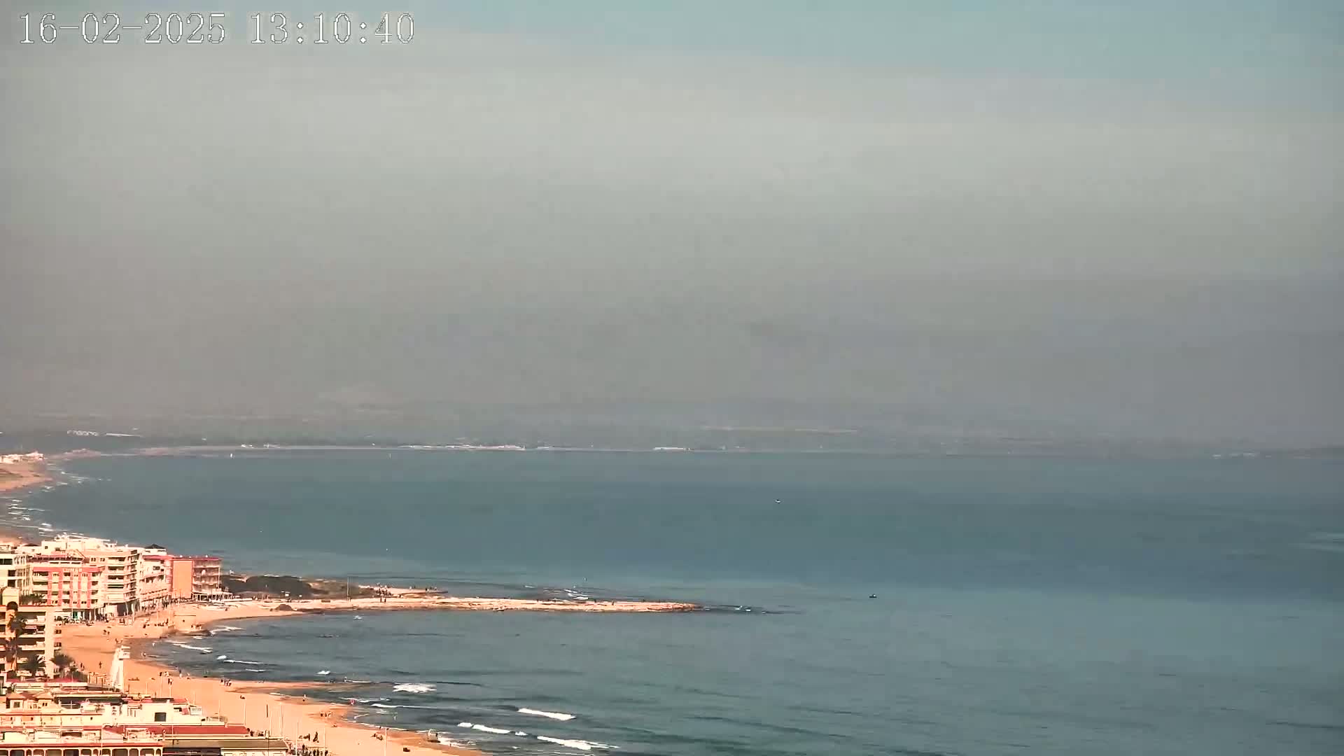 A hazy coastal scene shows a sandy beach with low buildings, calm ocean waves, and a distant hazy shoreline.