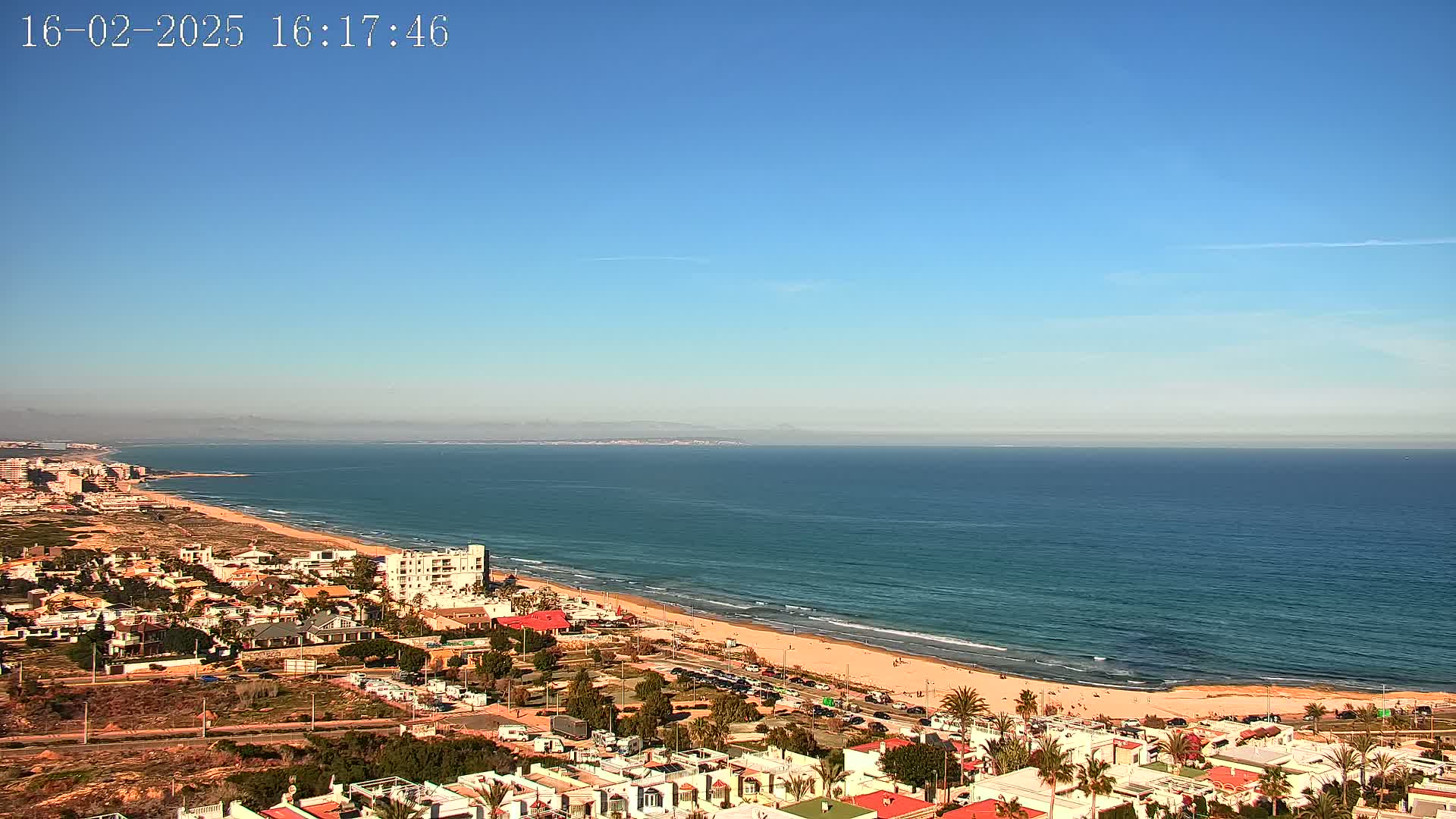 A coastal town overlooks a long sandy beach and calm ocean under a clear blue sky.