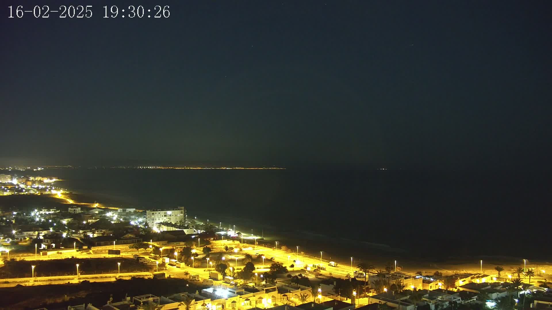 A nighttime coastal town is illuminated, with a dark sea extending to a distant, faintly lit shoreline under a clear, starry sky.