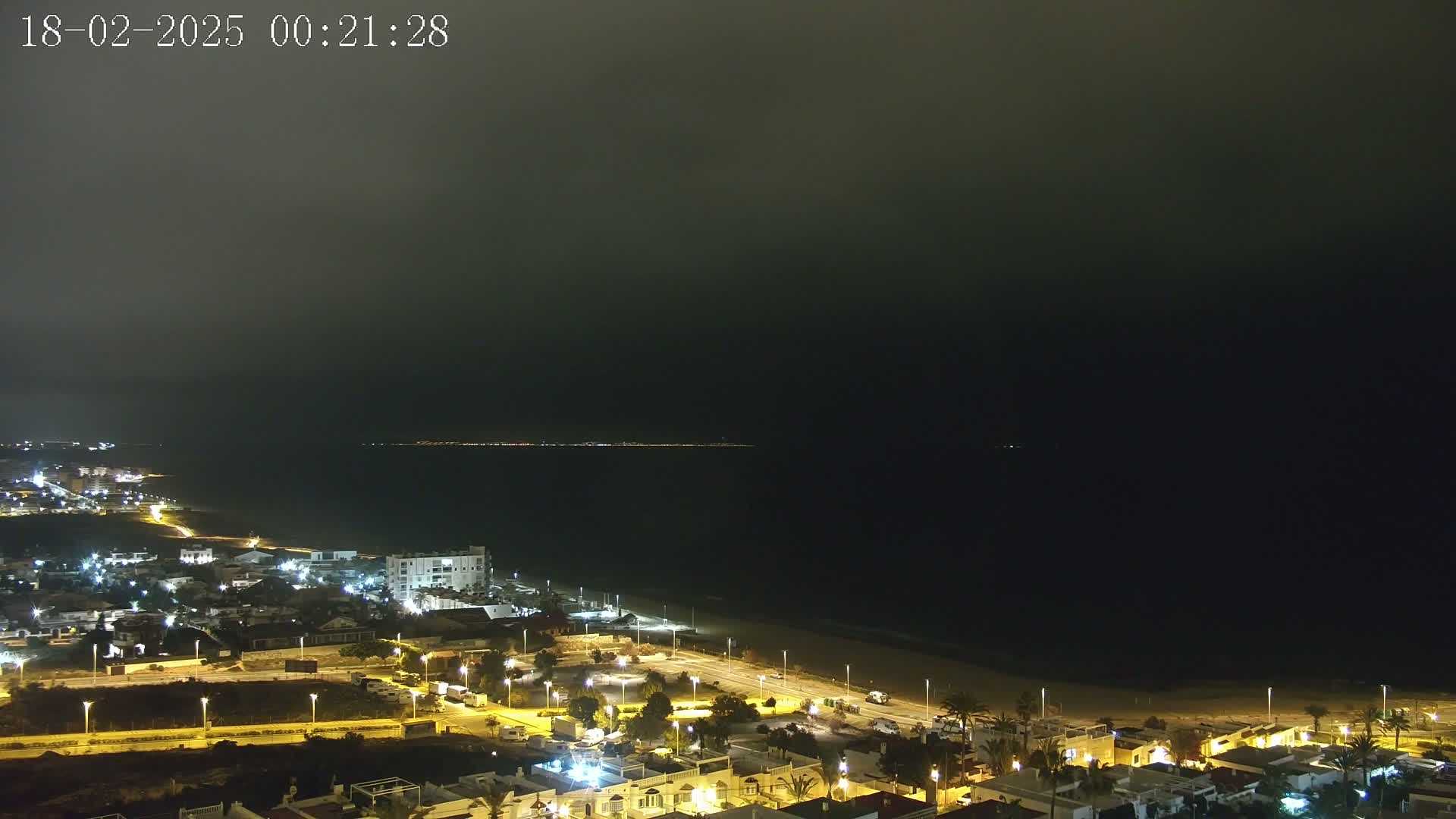 A nighttime coastal town with dimly lit buildings and streets overlooks a dark ocean under a hazy sky.