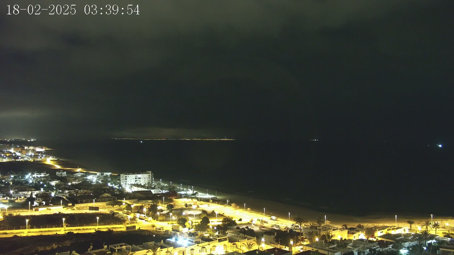 A nighttime coastal town with illuminated streets and buildings overlooks a dark ocean under a cloudy sky.
