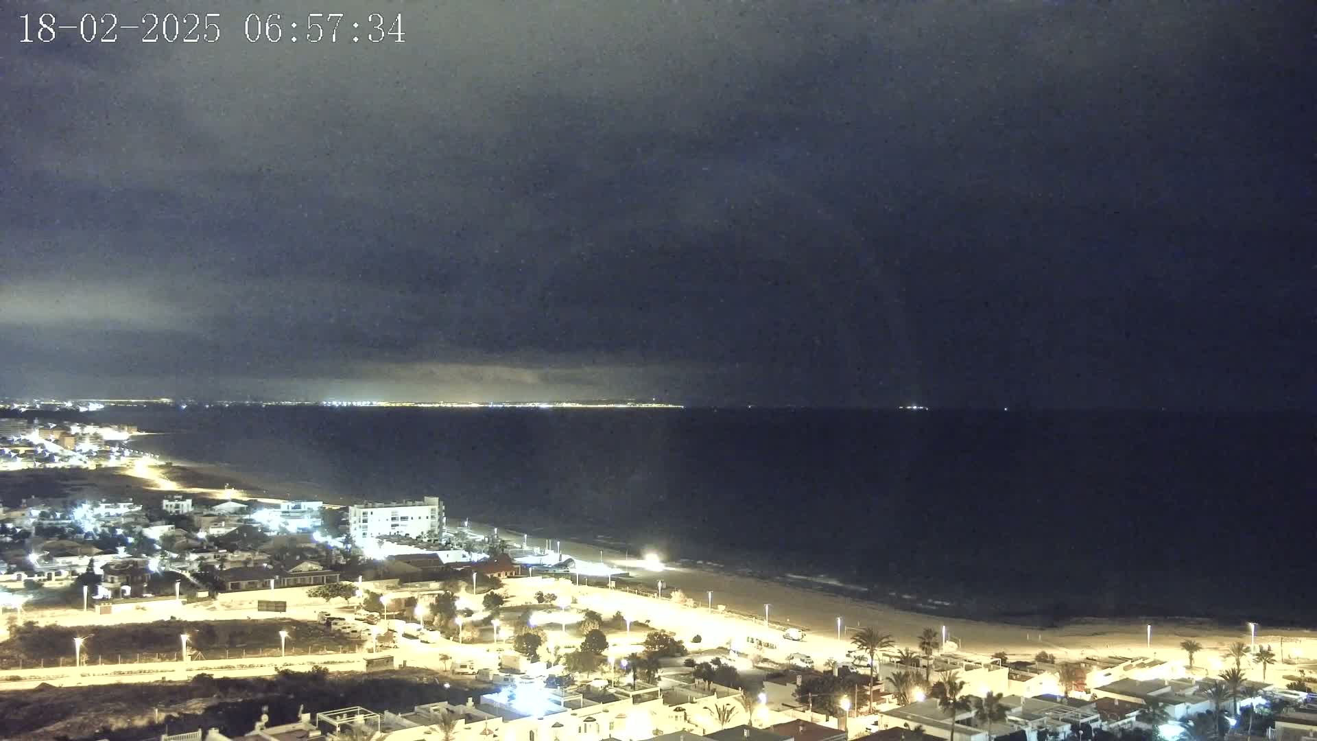 A nighttime coastal scene shows a dark, cloudy sky over a calm sea, with a lit-up beach and residential area along the shore.