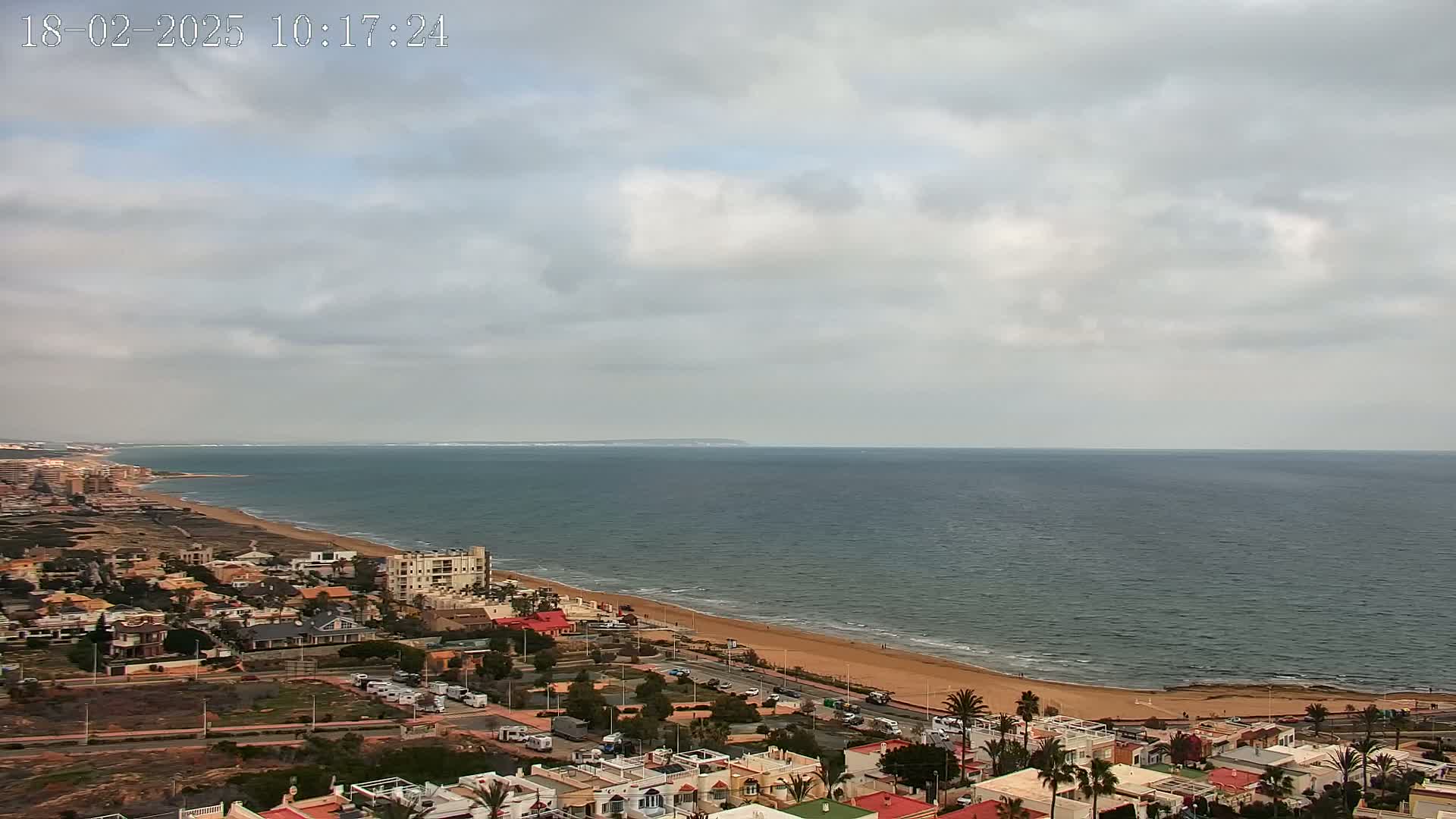 An overcast sky overlooks a sandy beach and coastal town beside a calm sea.