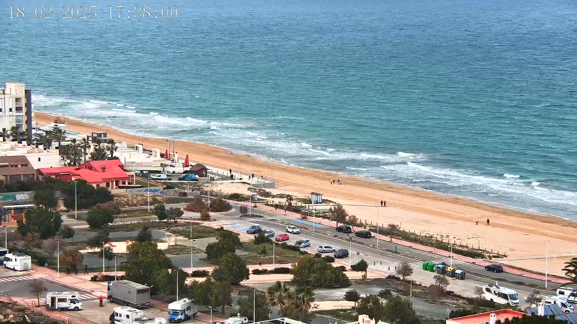 An aerial view shows a sandy beach with a few people walking along it, bordered by a road with parked cars and RVs, with a calm sea under a cloudy sky.
