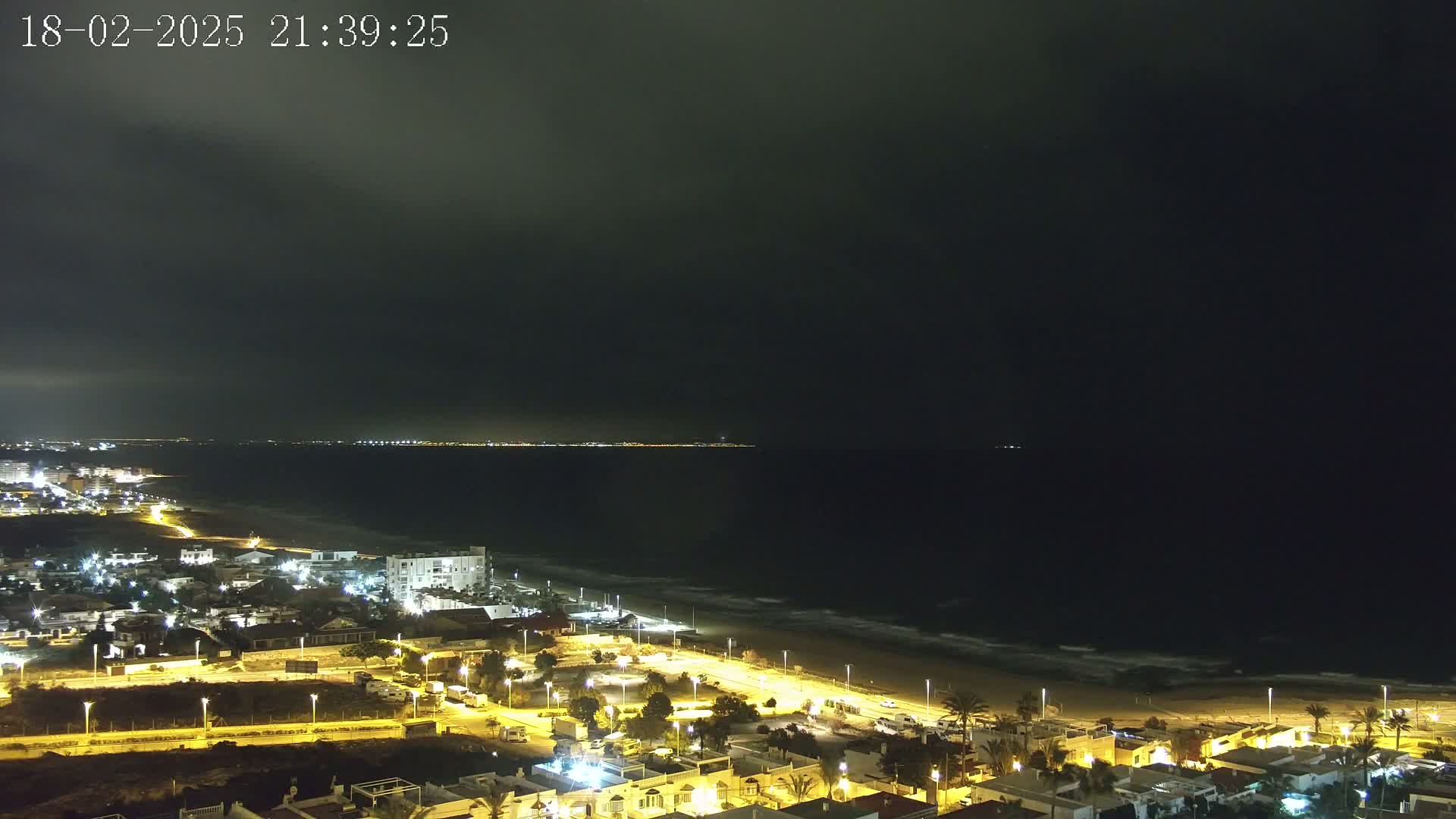 A nighttime coastal scene shows a dark ocean with small waves lapping a sandy beach, under a mostly cloudy sky, and a town illuminated with streetlights.