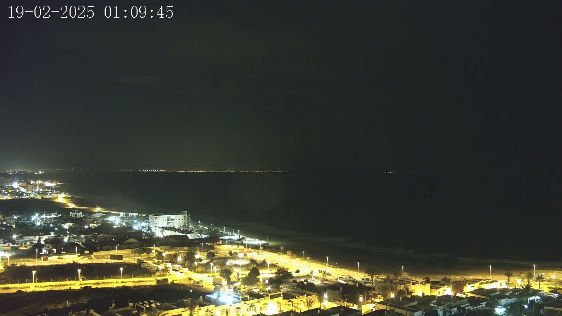 A coastal town at night, illuminated by streetlights, is seen overlooking a dark ocean with a distant shoreline faintly visible under a clear, dark sky.