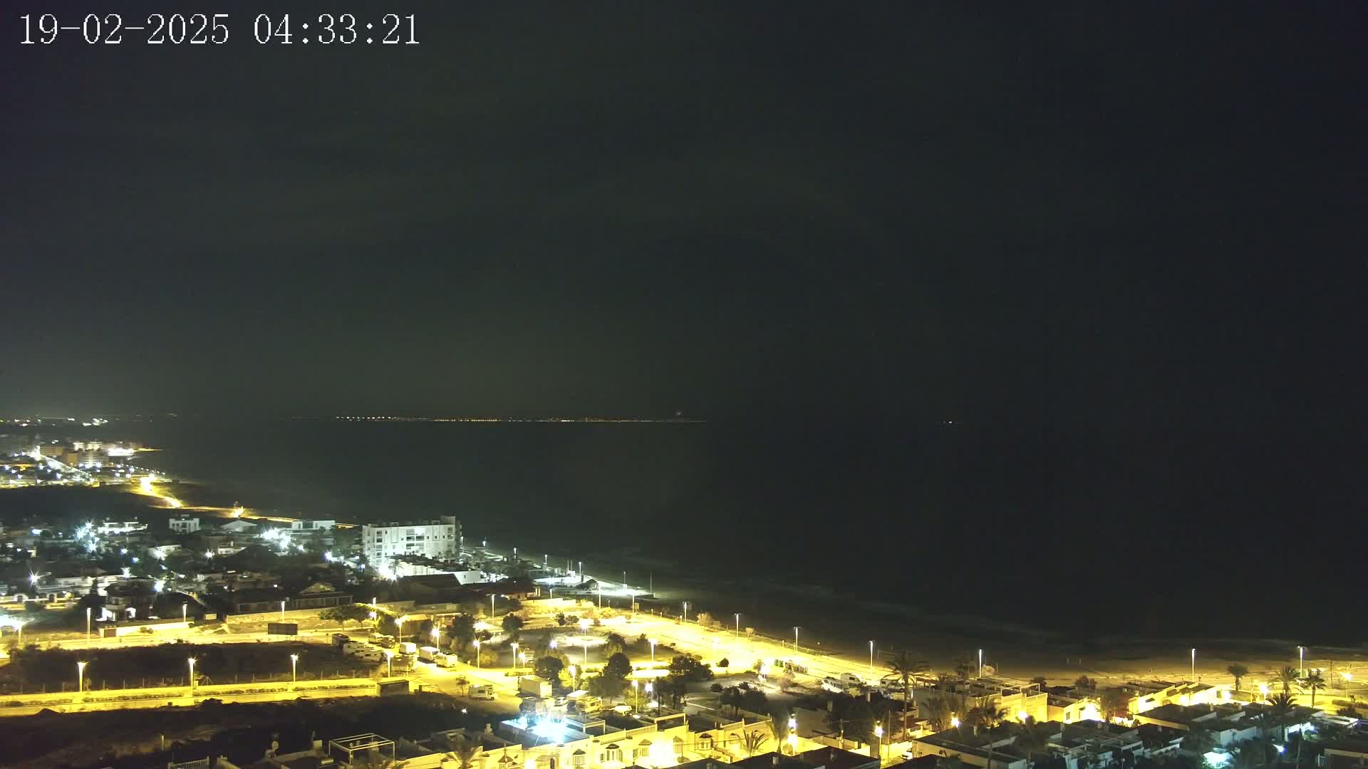 A nighttime coastal town illuminated by streetlights, with a dark ocean stretching to a dimly lit horizon.
