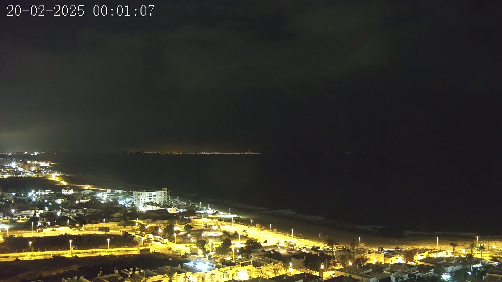 A nighttime coastal town illuminated by streetlights overlooks a dark ocean under a mostly cloudy sky.