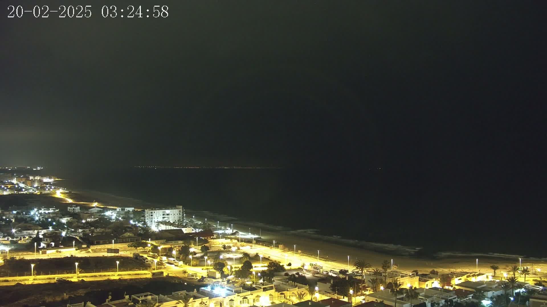 A nighttime coastal scene shows a dark ocean meeting a sandy beach, lined with illuminated buildings and streets.
