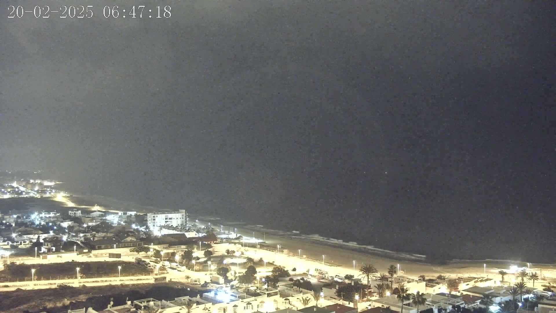 A nighttime coastal scene shows a town illuminated along a sandy beach with a dark, somewhat stormy ocean under a hazy sky.