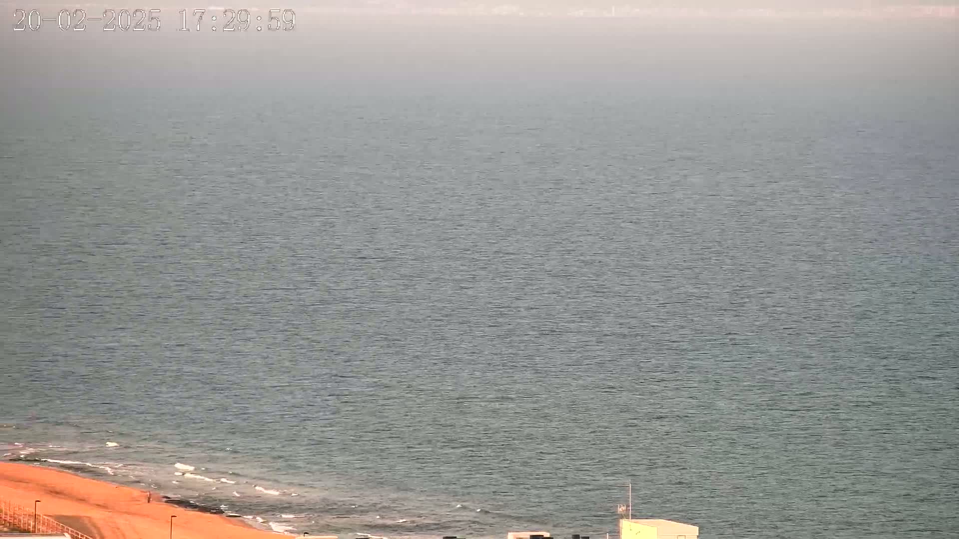 A hazy view of a calm sea meeting a sandy shore with a small building visible in the bottom right corner.