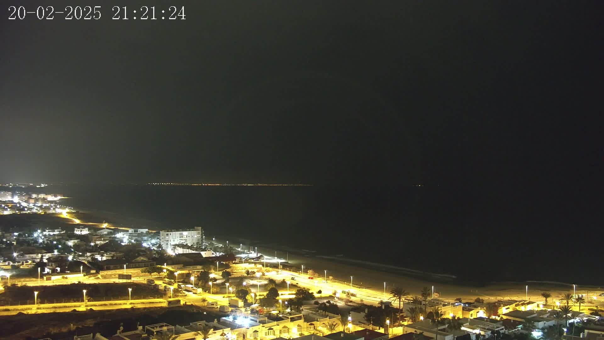 A nighttime coastal scene shows a dark ocean with small waves lapping a sandy beach, beside a town with illuminated streets and buildings.