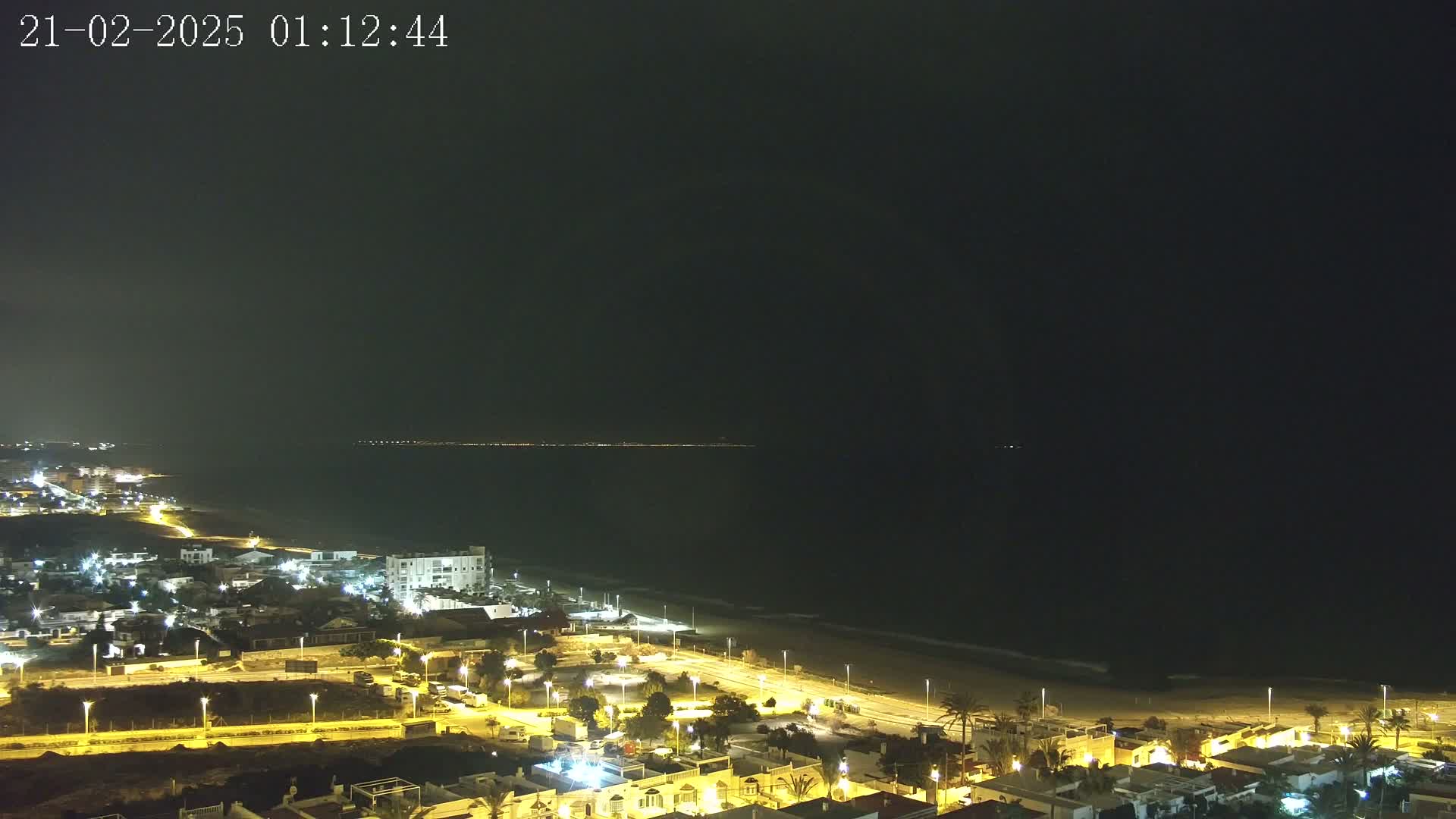 A nighttime coastal town is illuminated, overlooking a dark ocean with a faint light horizon under a clear, dark sky.
