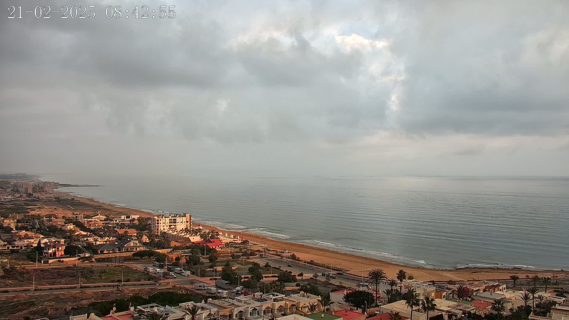An overcast sky hangs over a calm ocean meeting a sandy beach lined with coastal residential buildings.