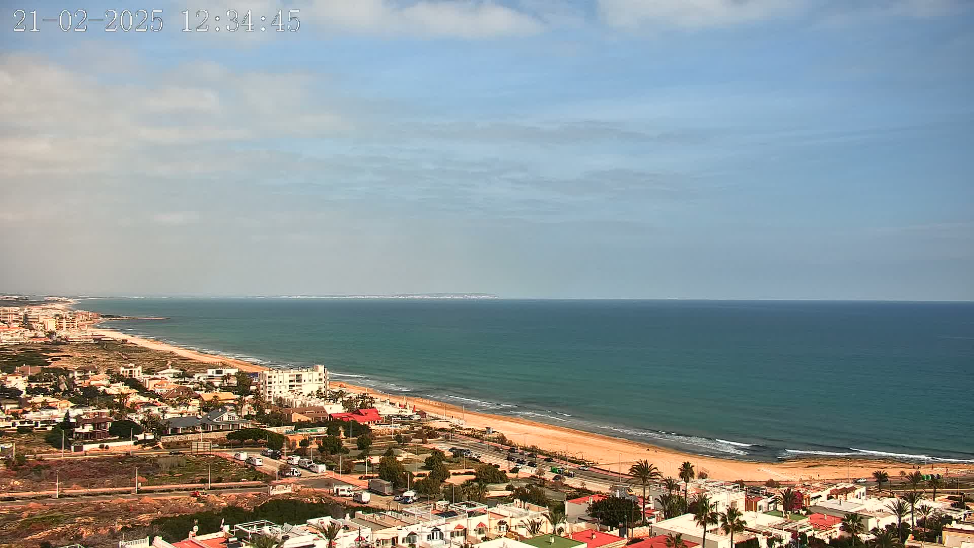 A coastal town overlooks a long sandy beach and a calm, teal ocean under a mostly clear sky with some scattered clouds.