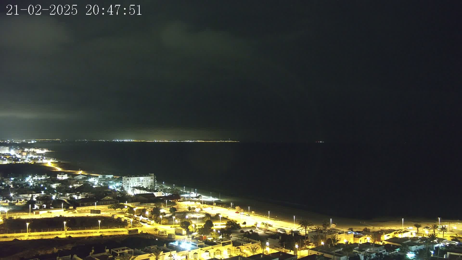A nighttime coastal scene shows a dark ocean, a sandy beach, and a town illuminated by streetlights under a mostly cloudy sky.