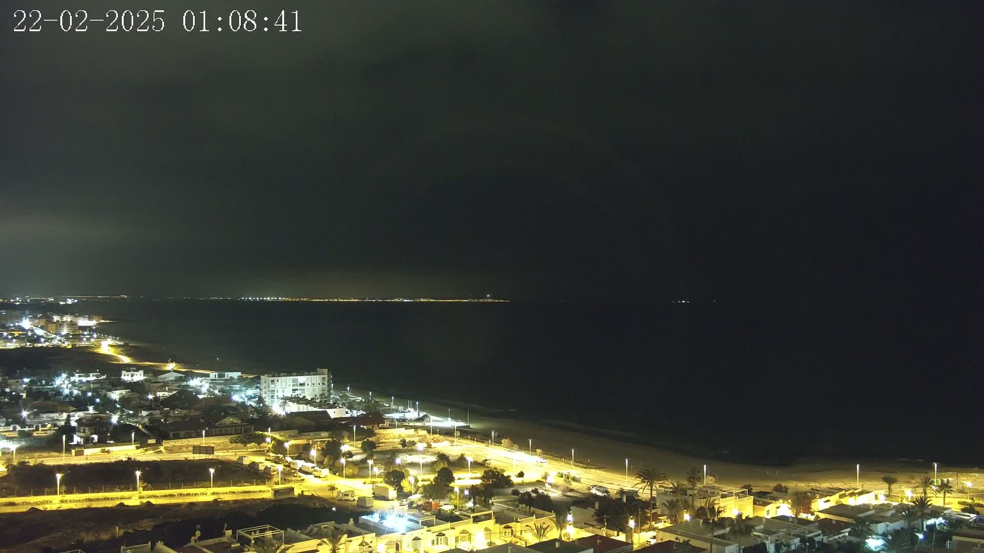 A coastal town at night is illuminated under a dark, cloudy sky, with a beach separating the town from a calm, dark ocean.