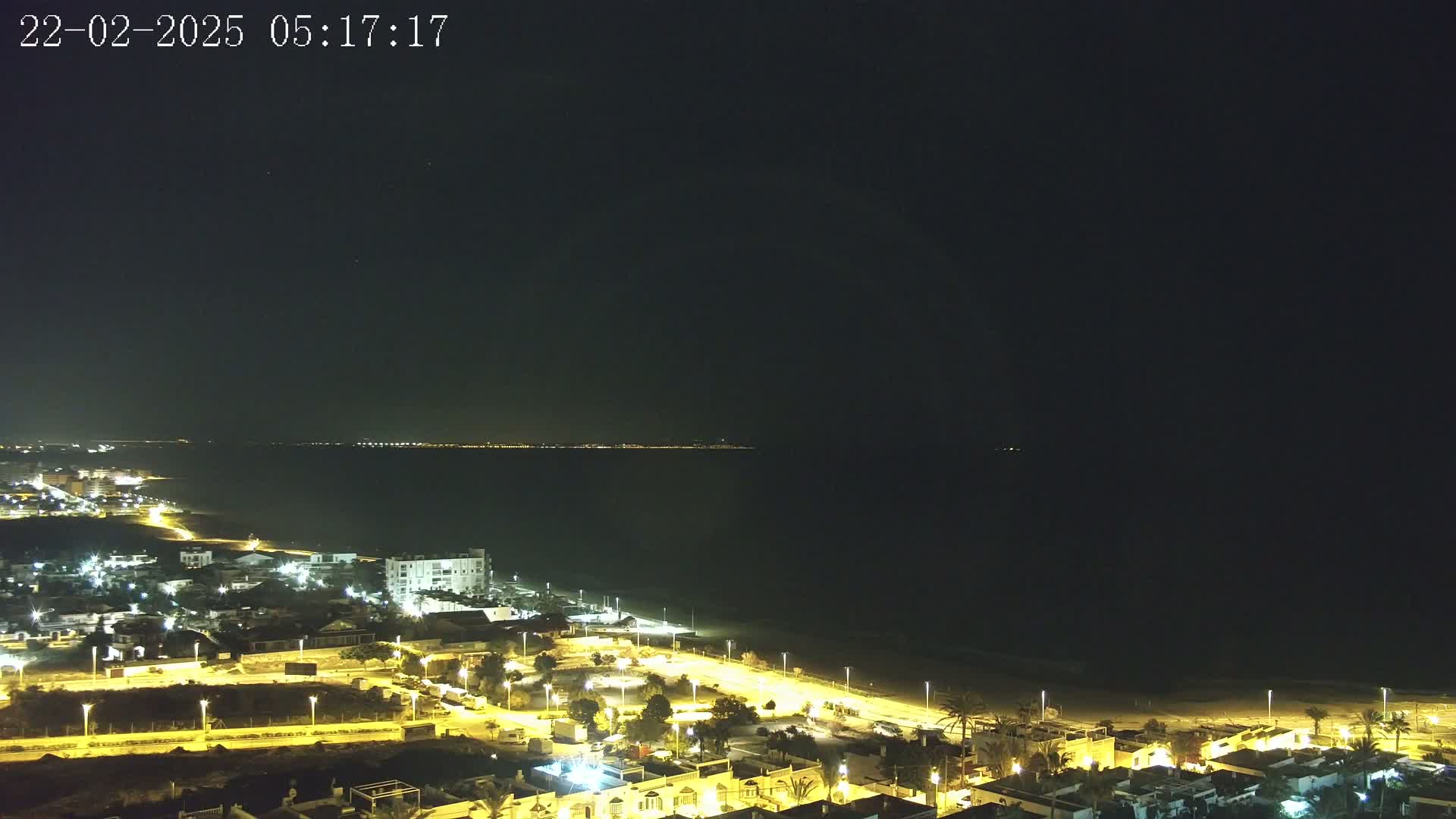 A nighttime coastal town illuminated by streetlights is visible along a dark, calm ocean under a clear night sky.