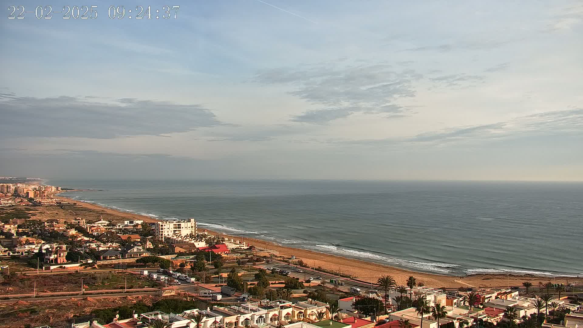 An aerial view shows a coastal town under a partly cloudy sky, with a calm ocean meeting a sandy beach.