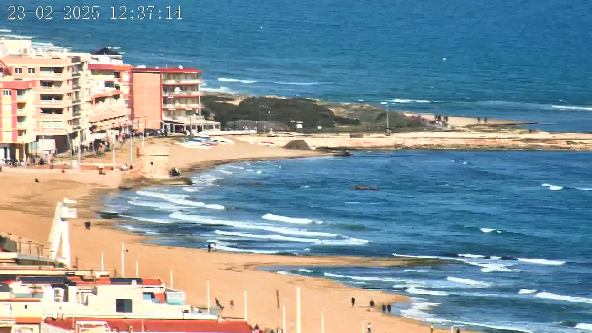 A sandy beach with moderate waves laps against the shore, near buildings and a small stone tower under a sunny sky.