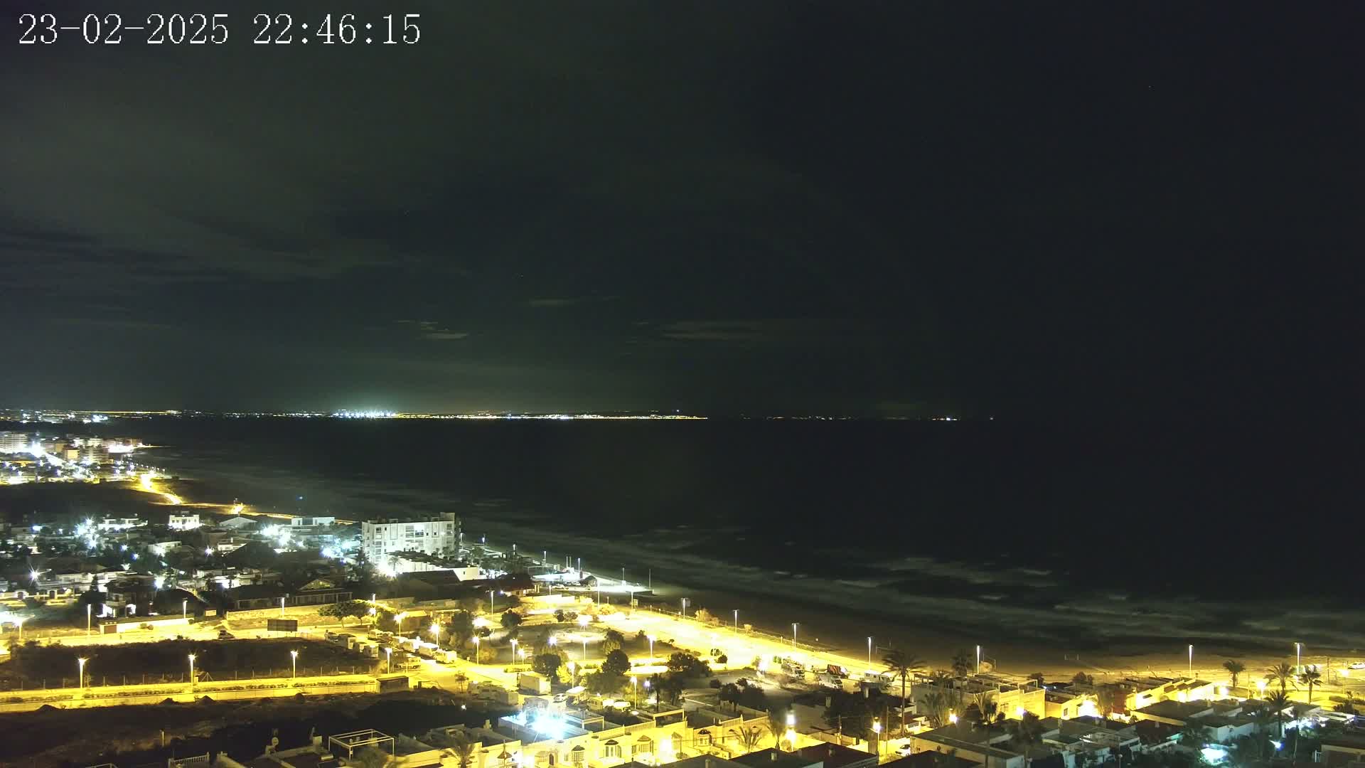 A nighttime aerial view shows a coastline with a town illuminated by streetlights, a dark ocean with small waves, and a distant city skyline under a mostly clear, dark sky.