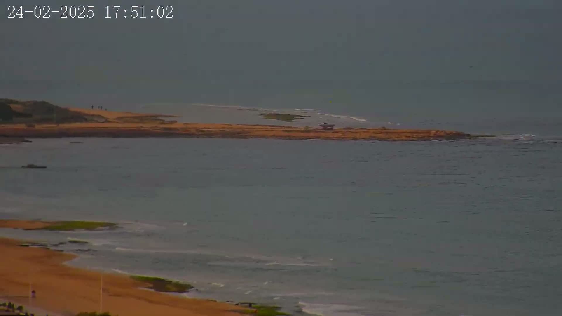 A sandy beach and calm, overcast ocean are visible from a high vantage point, with a small structure on a nearby landmass.