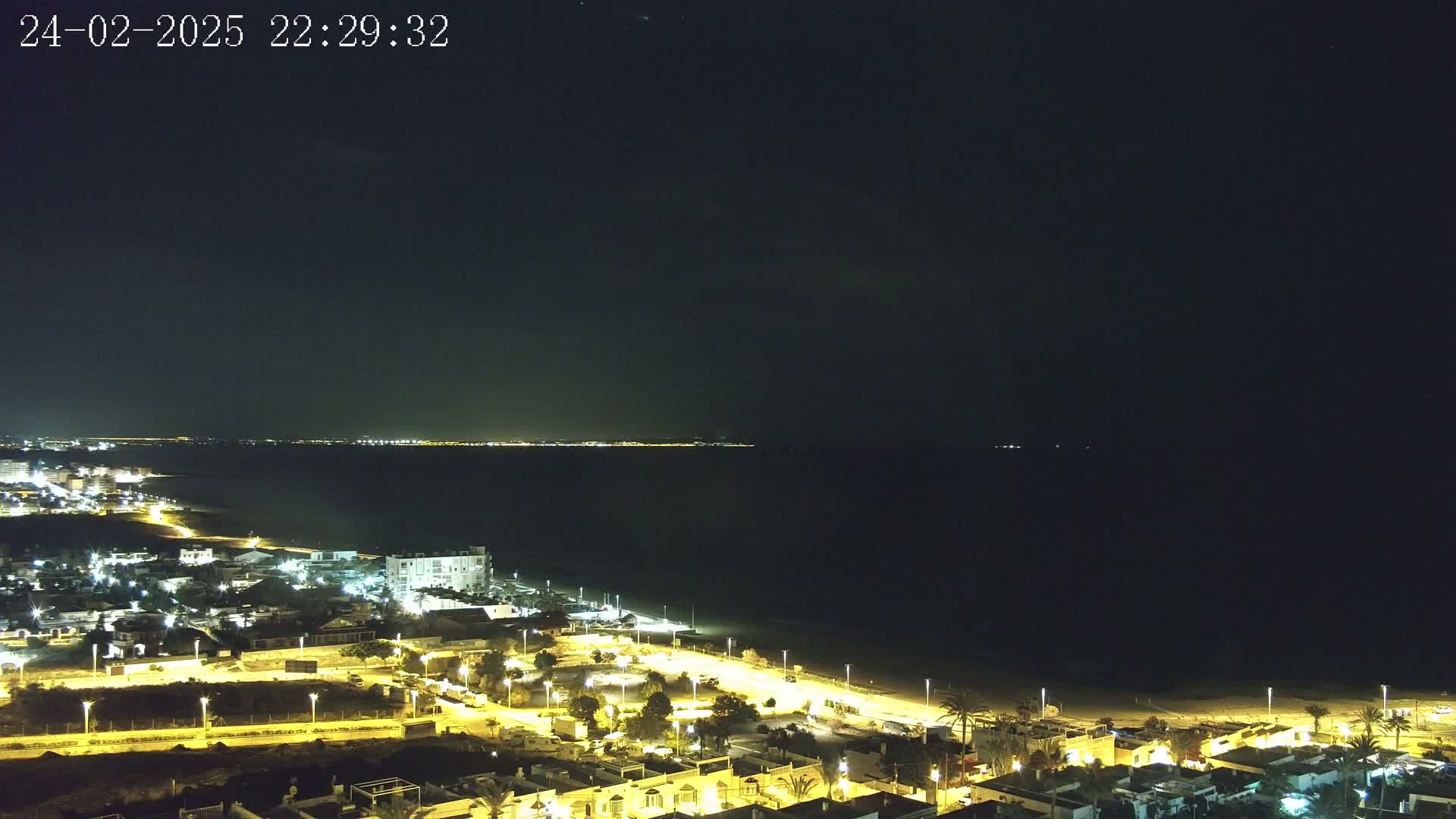 A nighttime coastal town is illuminated by streetlights, overlooking a dark ocean with a distant, faintly lit shoreline under a clear, starlit sky.