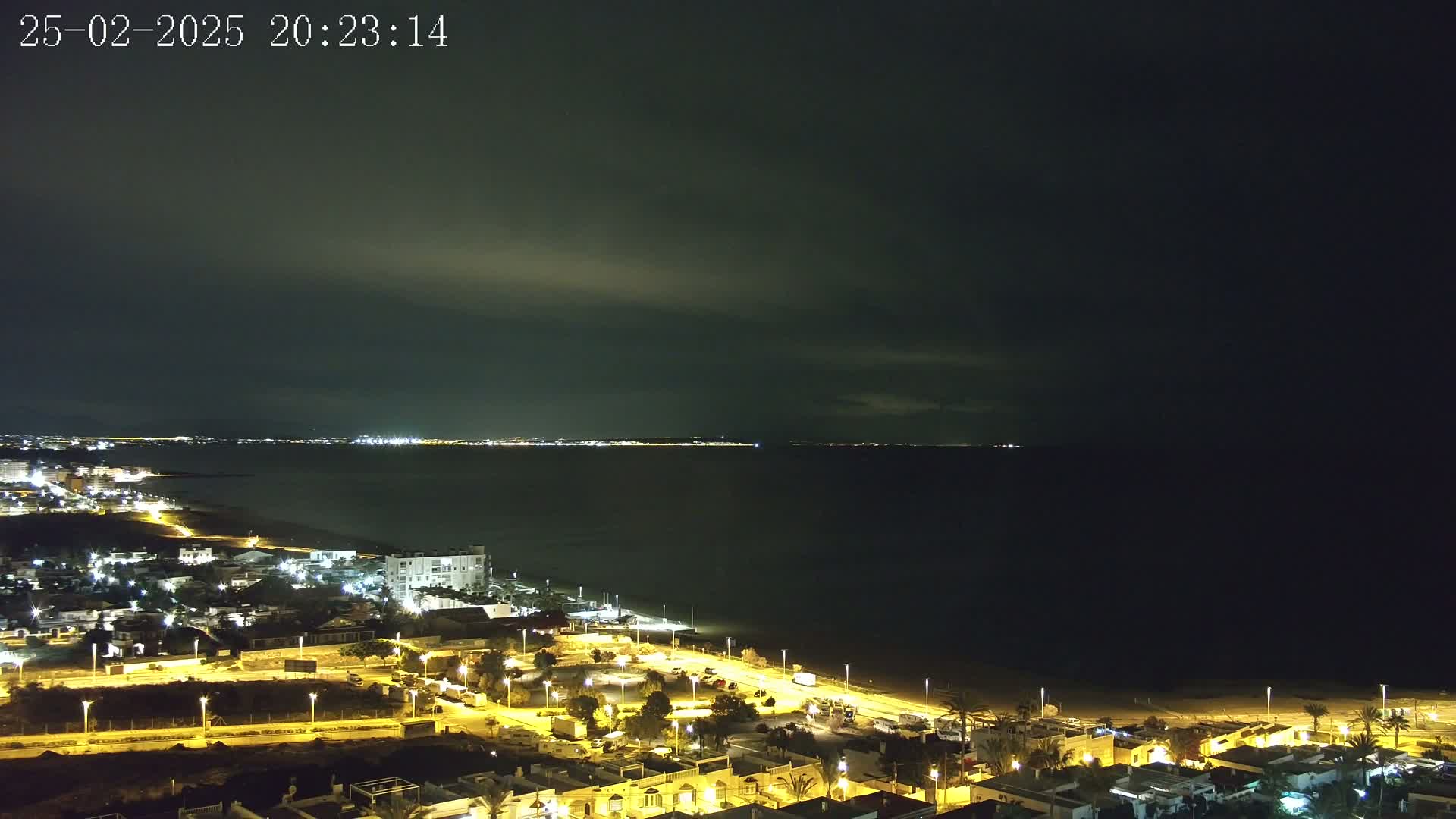 A nighttime coastal view shows a dark sea under a mostly cloudy sky, with a line of illuminated buildings along the shore and more lights visible in the distance across the water.