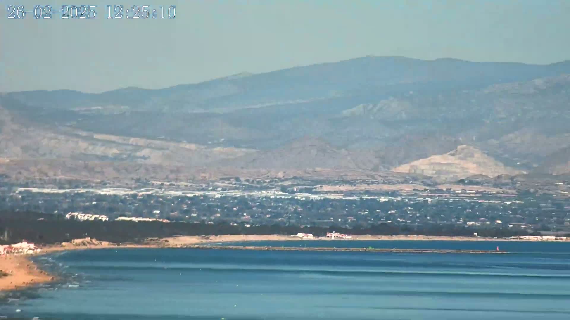A coastal scene shows a sandy beach, calm ocean, a town nestled behind a line of trees, and hazy mountains in the background under a clear sky.