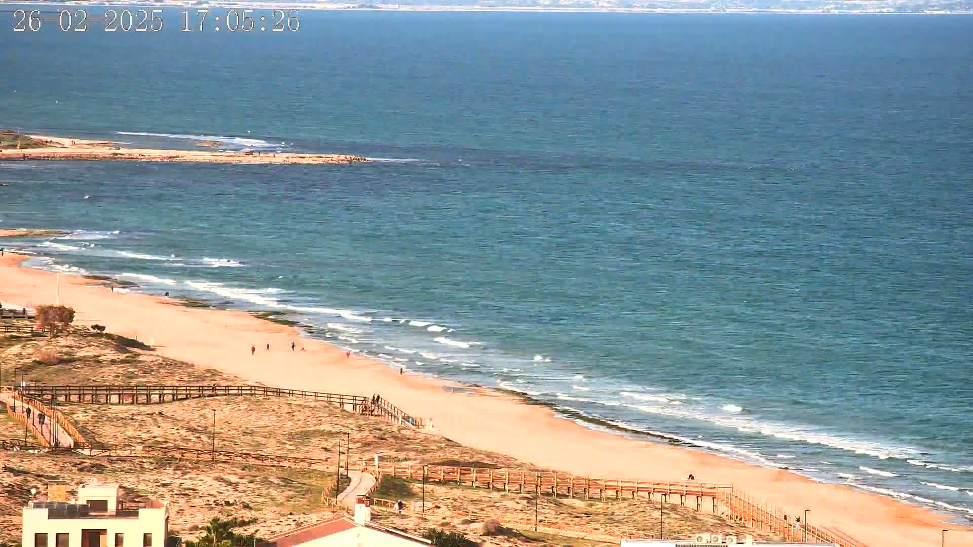 A sandy beach with a few people walking along the shore and wooden walkways, meets a teal ocean with small waves on a sunny day.